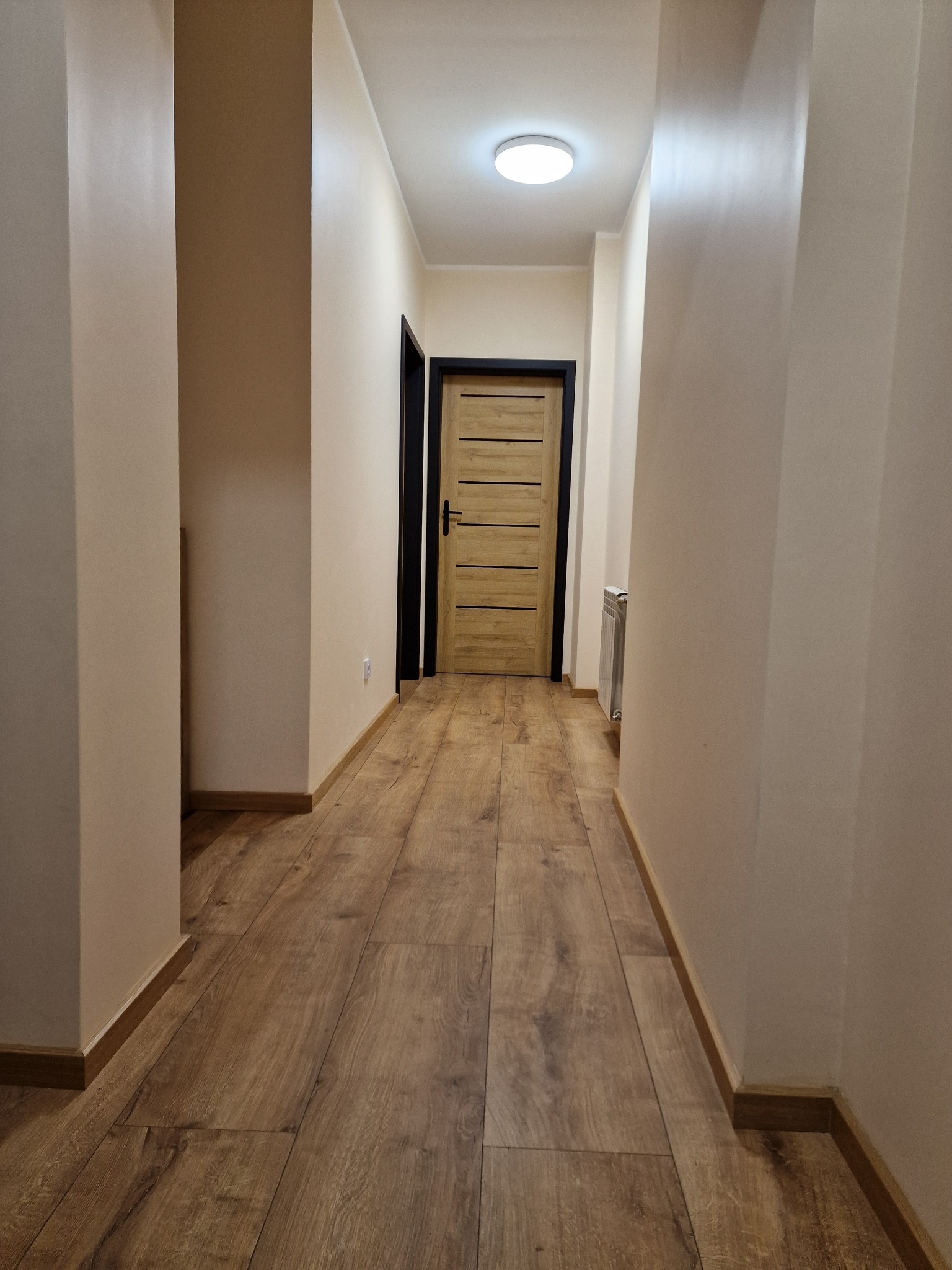 Korytarz po remoncie z drewnopodobną podłogą, świeżo pomalowanymi ścianami w odcieniach beżu i nowymi drzwiami z czarną ościeżnicą oraz grzejnikiem.
