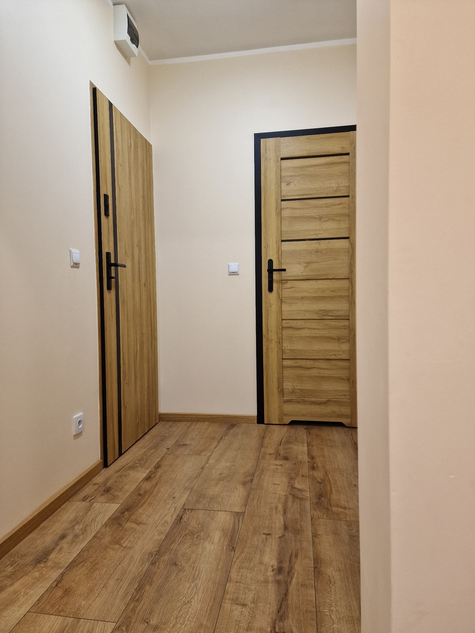 Korytarz z dwiema drewnianymi drzwiami z czarnymi obramowaniami i klamkami, parkiet na podłodze, jasne ściany, widoczna instalacja elektryczna.