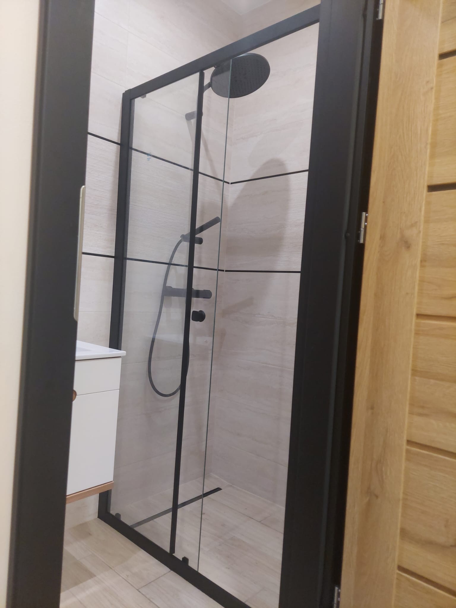 Nowoczesna kabina prysznicowa z czarnymi profilami i deszczownicą, widok z boku na umywalkę i fragment drewnianej okładziny ściany.