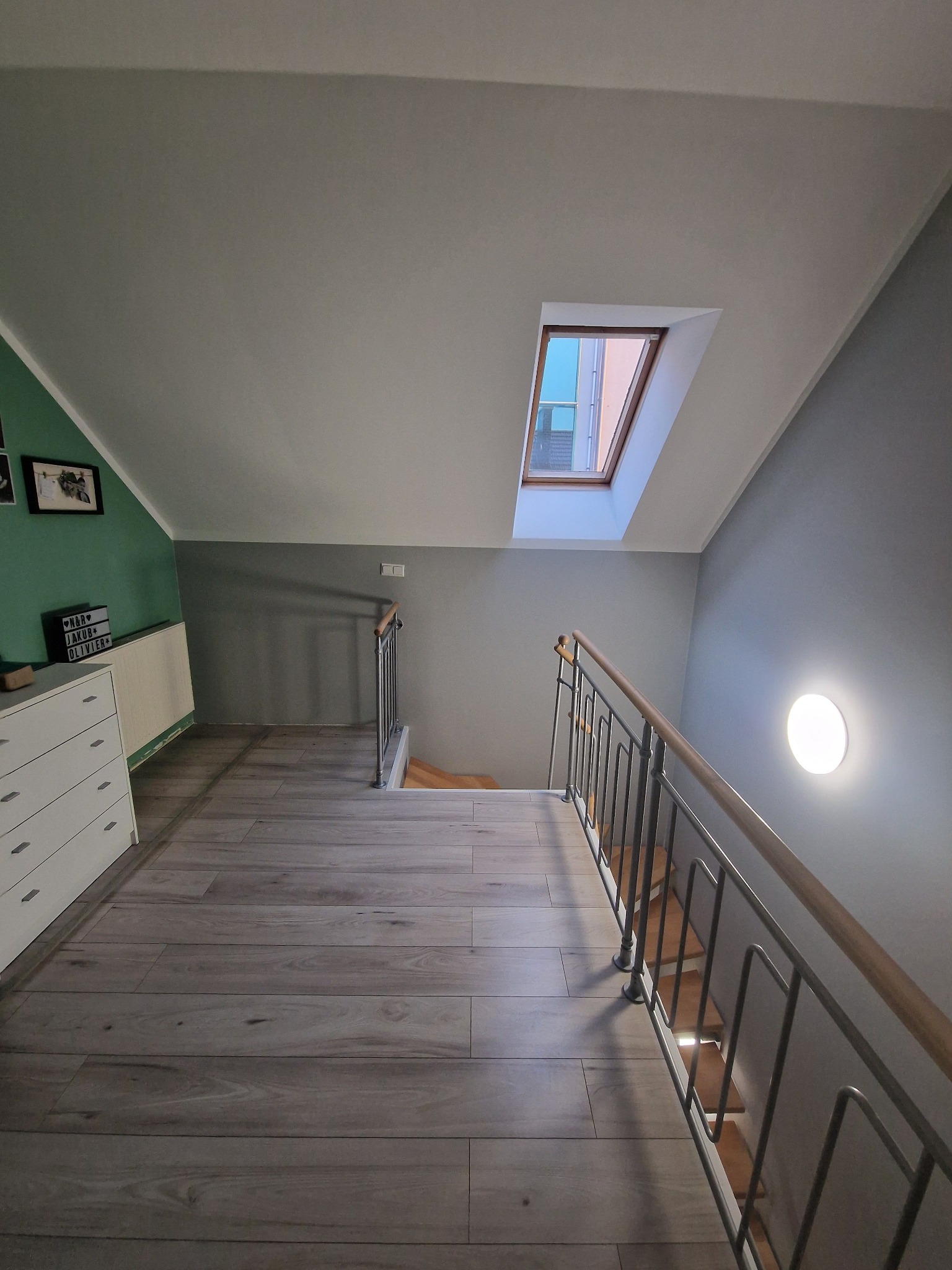 Poddasze z drewnianą podłogą, schodami z metalową balustradą i oknem dachowym, ściany pomalowane na szaro i zielono.