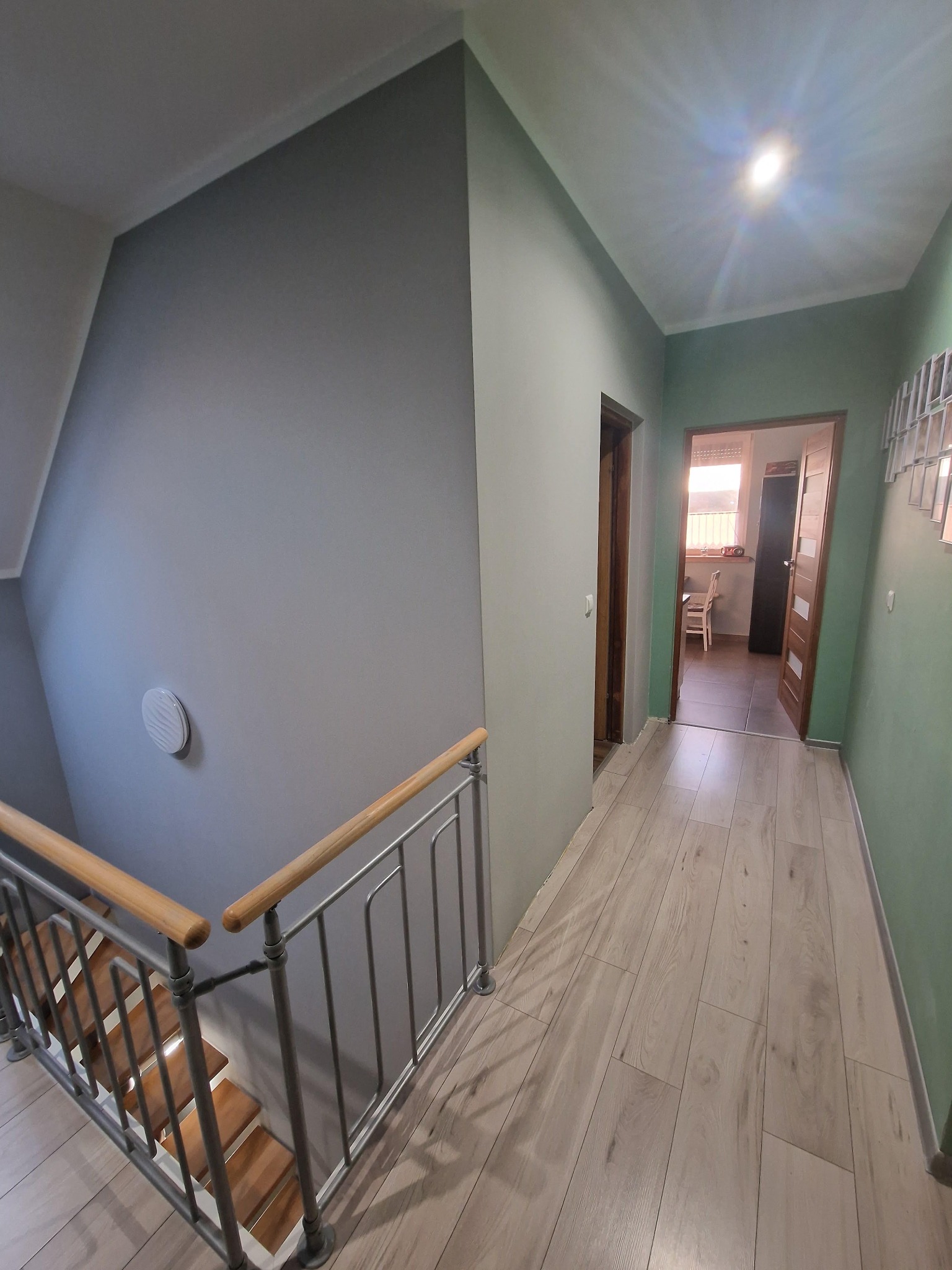 Korytarz po odświeżeniu: ściany pomalowane na szaro i zielono, drewniane schody z metalową barierką, panele podłogowe imitujące jasne drewno, widok na otwarte drzwi do pokoju z jasnymi meblami.