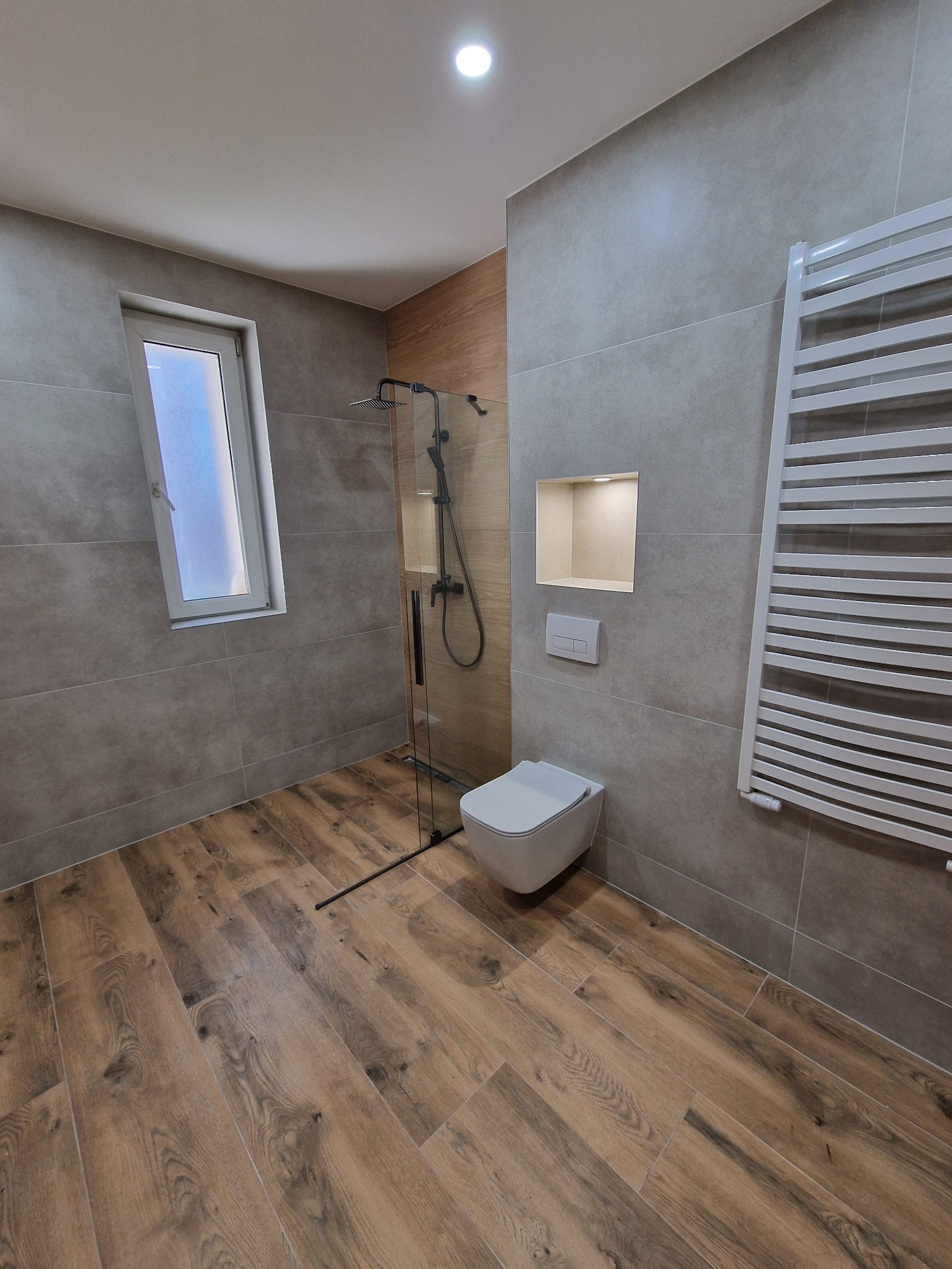 Nowoczesna łazienka po remoncie: szare płytki, drewnopodobna podłoga, kabina prysznicowa ze szkła, wisząca toaleta i grzejnik drabinkowy. Oświetlona wnęka ścienna.