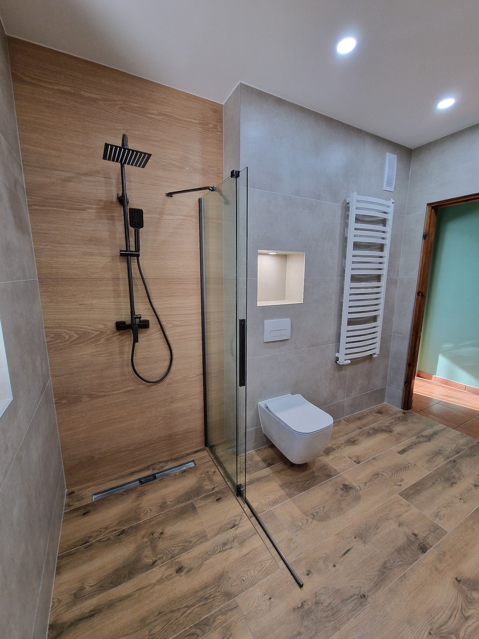 Nowoczesna łazienka po remoncie: kabina walk-in z czarną armaturą, drewnopodobne płytki, grzejnik drabinkowy i podtynkowa spłuczka.