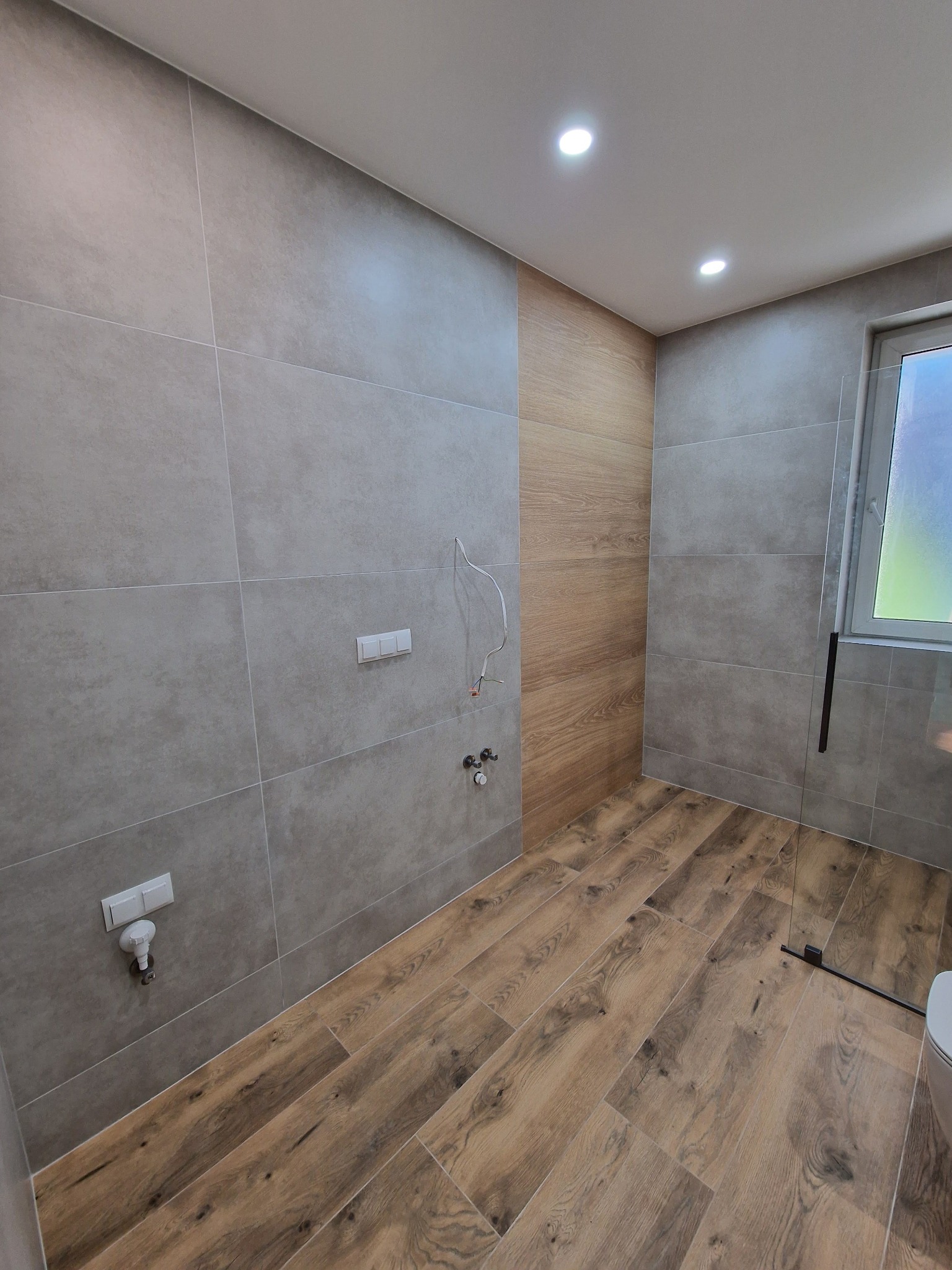 Wykończona łazienka z szarymi płytkami ściennymi i drewnopodobną podłogą. Widoczne punkty instalacyjne, szklana kabina prysznicowa i okno.