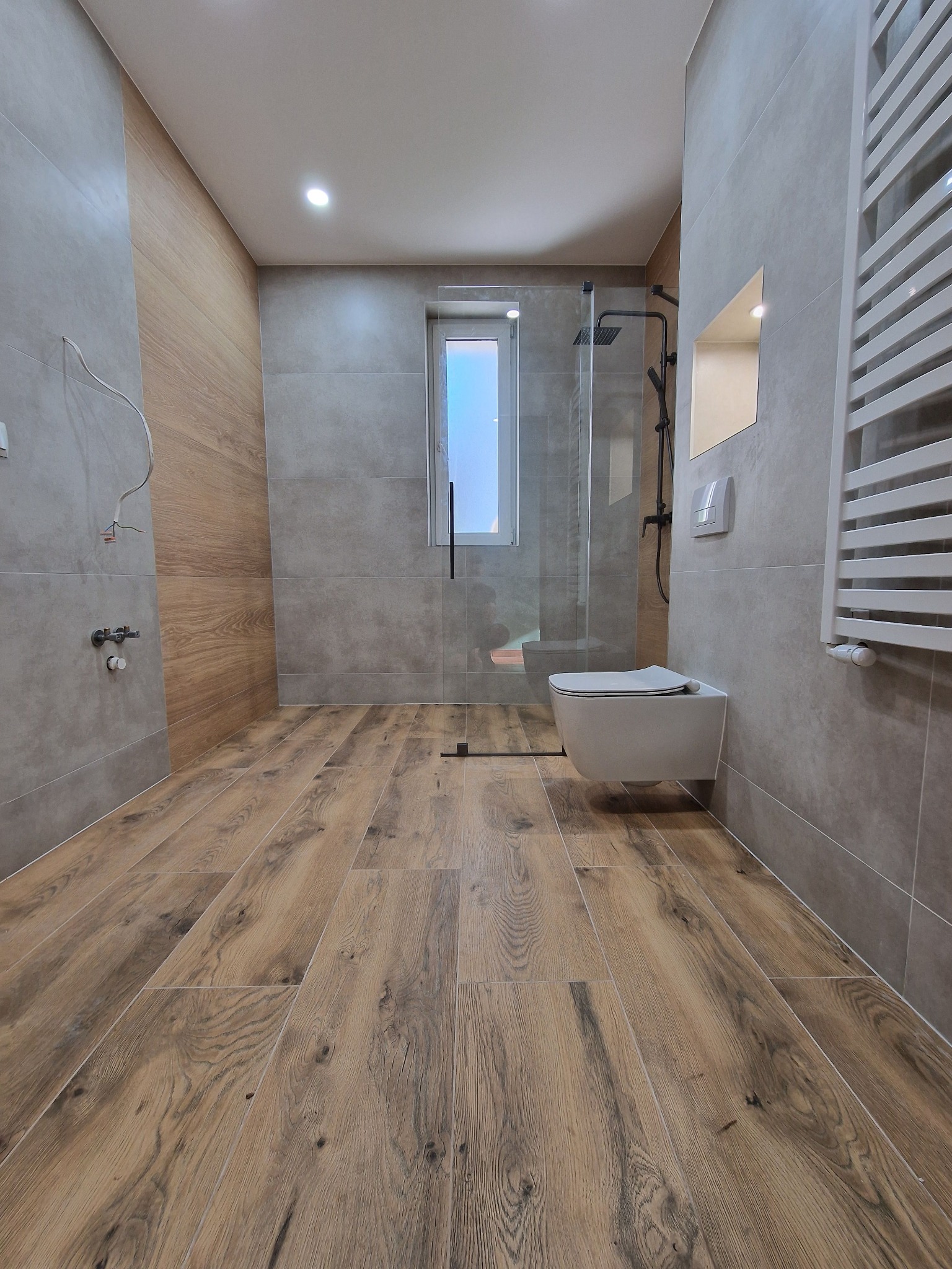 Nowoczesna łazienka z drewnopodobną podłogą, szarymi płytkami i kabiną prysznicową typu walk-in. Widoczne podłączenia hydrauliczne i grzejnik drabinkowy.