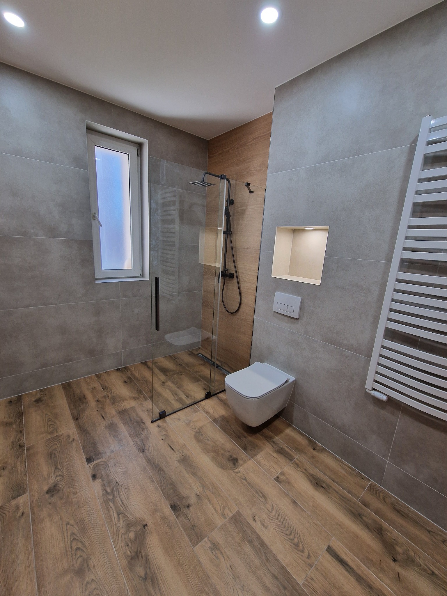 Nowoczesna łazienka z kabiną prysznicową typu walk-in, drewnopodobną podłogą i szarą płytką. Uwagę zwraca podświetlona wnęka w ścianie i minimalistyczna toaleta.