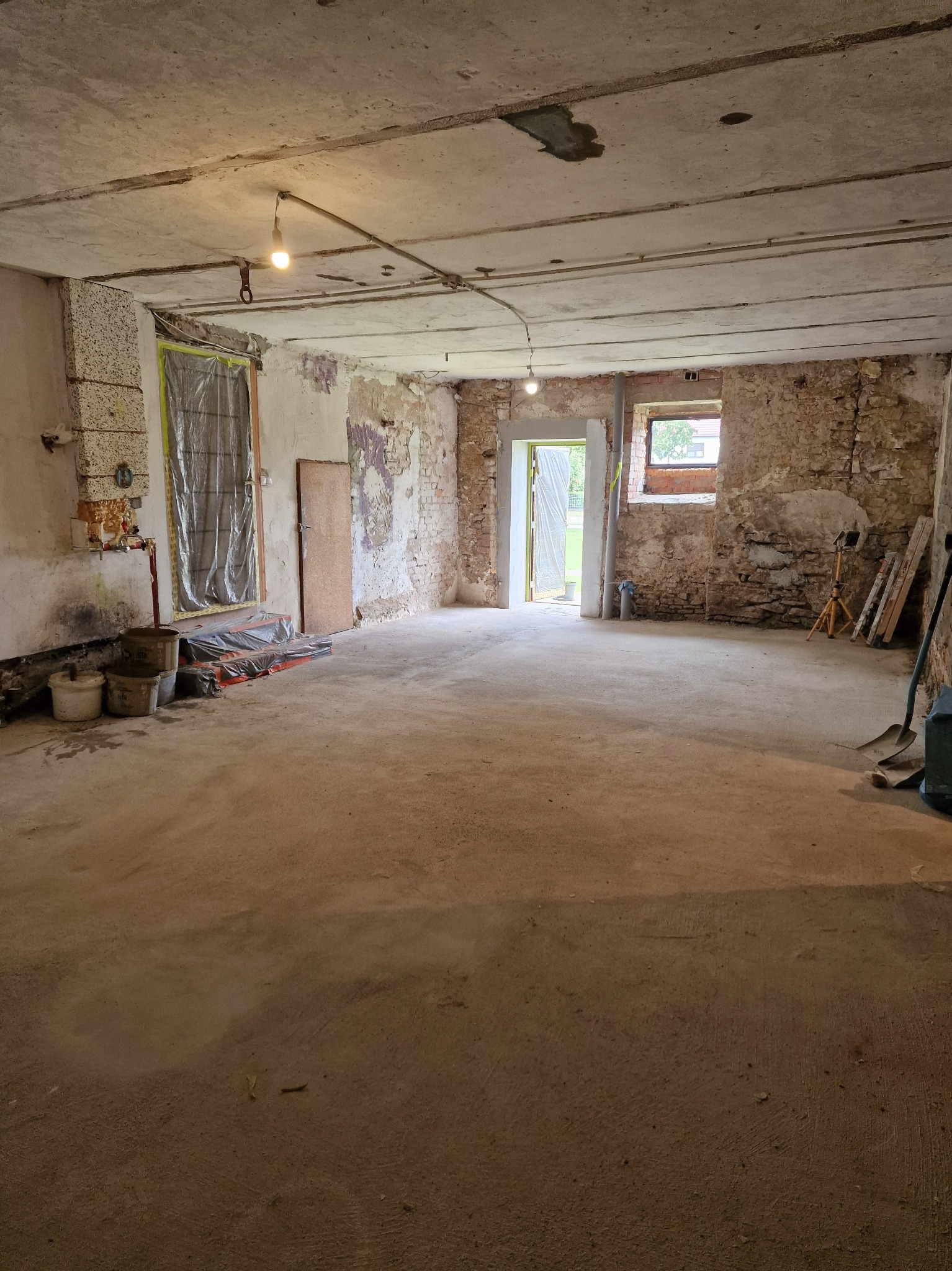 Wnętrze w trakcie prac remontowych: surowe ściany z cegły i betonu, zabezpieczone okna i drzwi folią, widoczne narzędzia i materiały budowlane. Przygotowanie do dalszych prac.