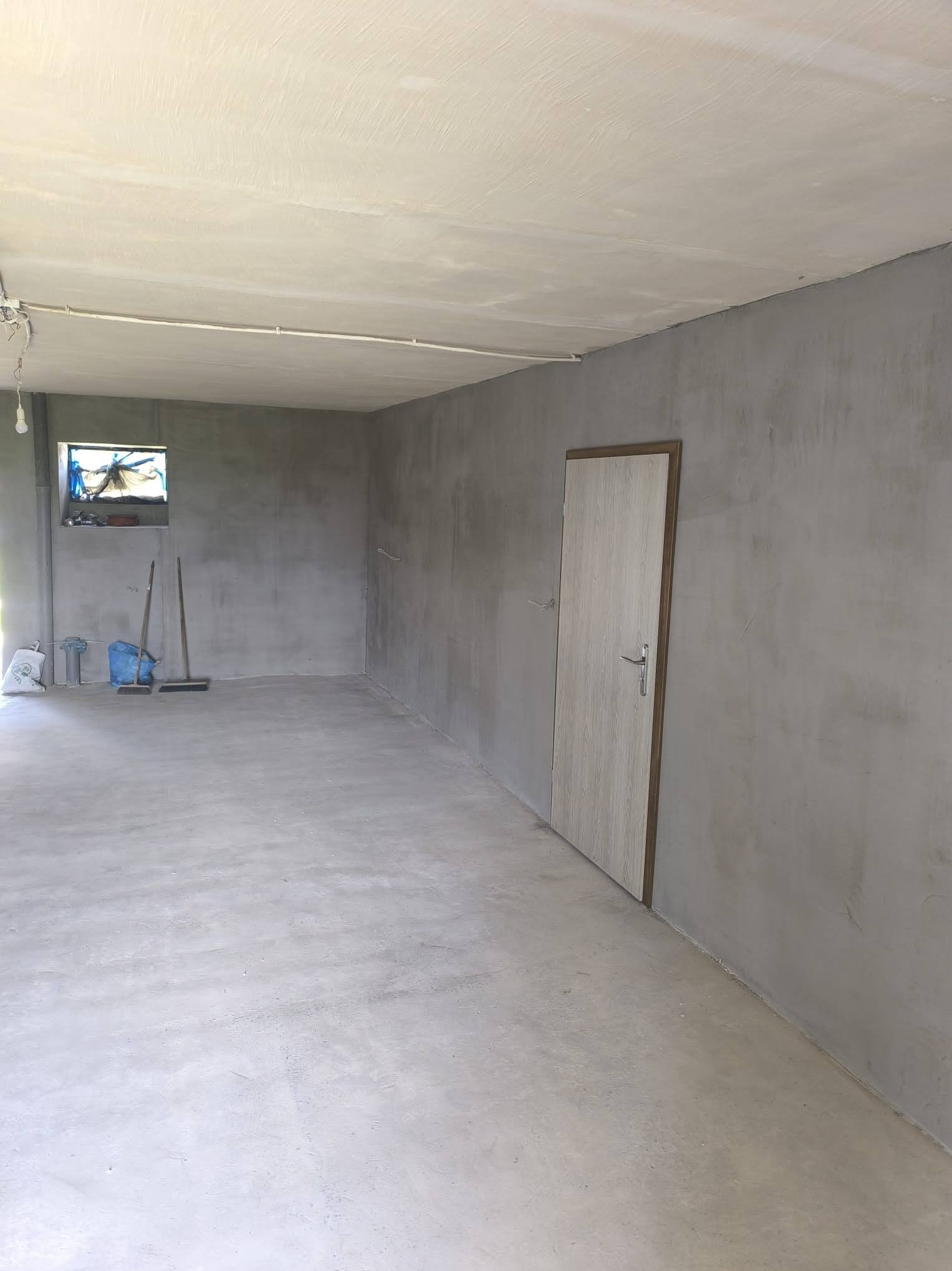 Szara, pusta przestrzeń po gładziowaniu ścian i wylewce podłogi, z widocznymi narzędziami i zamontowanymi drzwiami. Okno z lewej strony doświetla wnętrze.