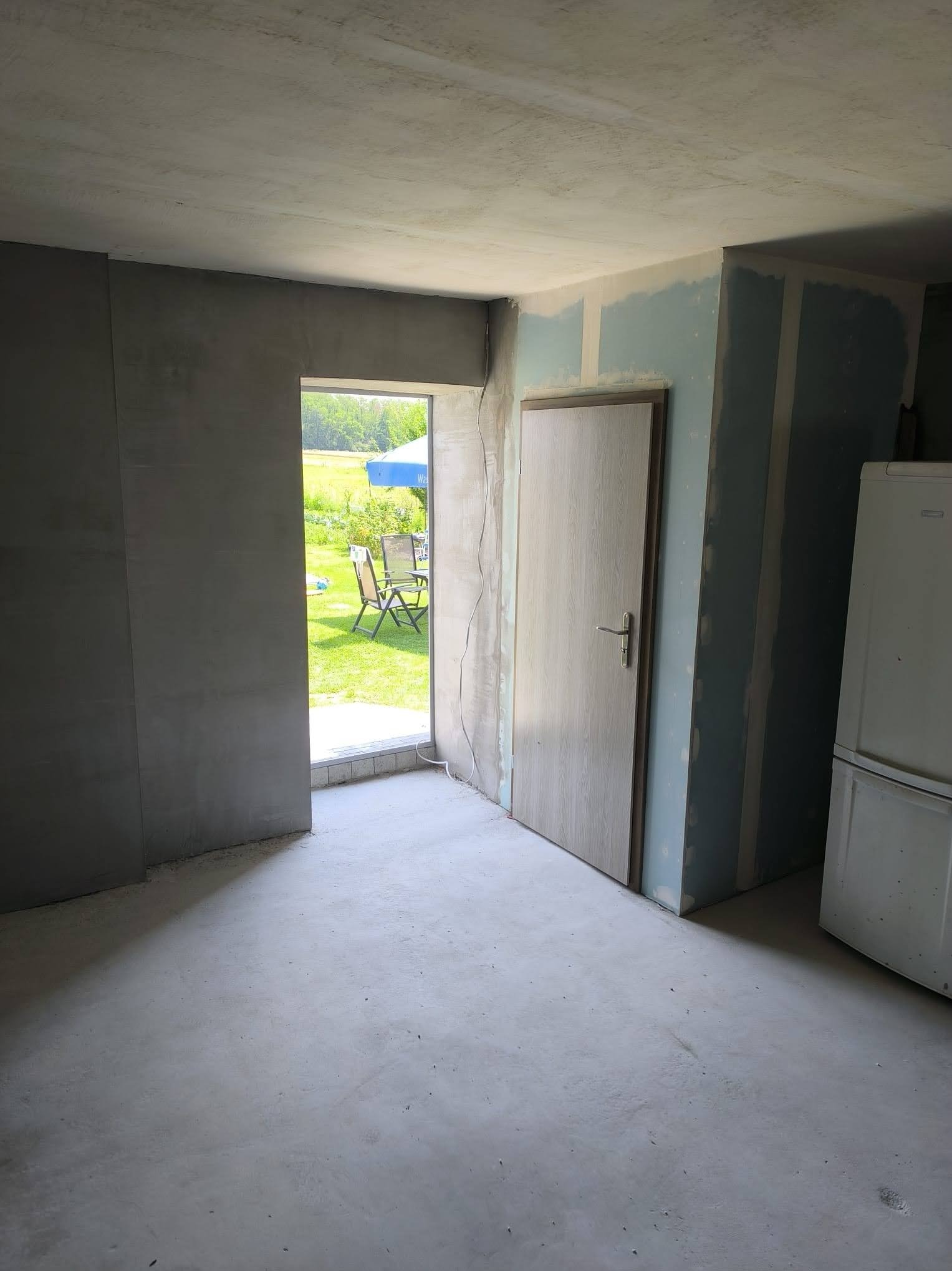 Wnętrze w trakcie prac wykończeniowych: ściany z gładzią, płyty karton-gips, osadzone drzwi, widoczny fragment lodówki. W tle widok na zewnątrz przez niezabezpieczony otwór.