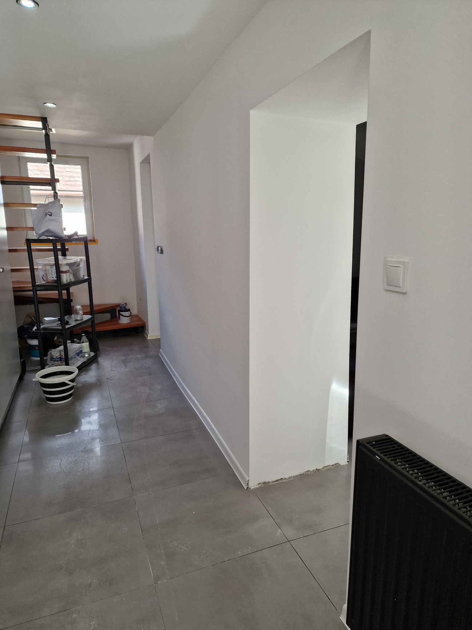 Świeżo pomalowane, białe ściany w korytarzu z szarą podłogą, widoczny grzejnik i fragment regału z akcesoriami malarskimi. Oświetlenie sufitowe.