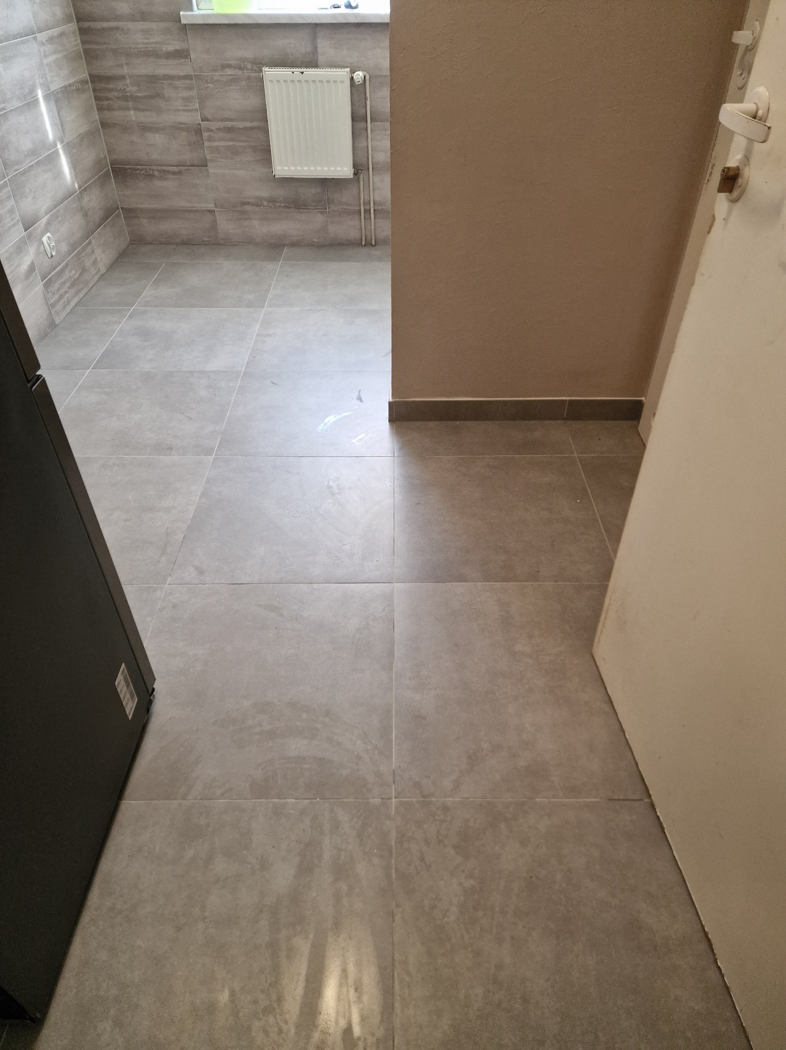 Wykończona łazienka z szarymi płytkami na podłodze i ścianach. Widoczny biały grzejnik na ścianie i fragment otwartych drzwi po prawej stronie. Minimalistyczny design.