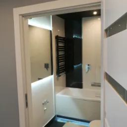 Nowoczesna łazienka z czarno-białymi płytkami, podświetlanym lustrem i grzejnikiem drabinkowym, widok zza uchylonych drzwi z widocznym fragmentem niezabezpieczonej podłogi.