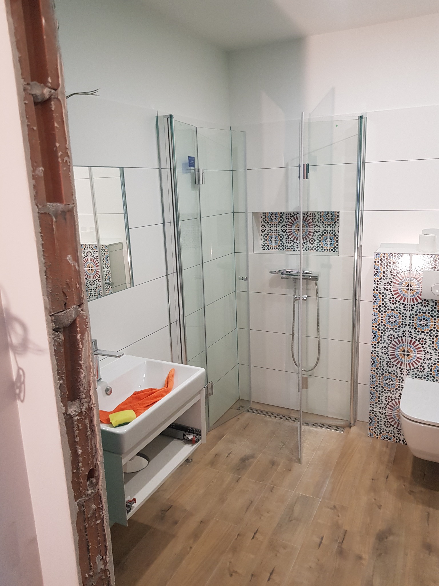 Łazienka w trakcie remontu: widoczna niedokończona ościeżnica, kabina prysznicowa z mozaiką, umywalka z pomarańczowym ręcznikiem i żółtą gąbką, drewnopodobne płytki podłogowe.