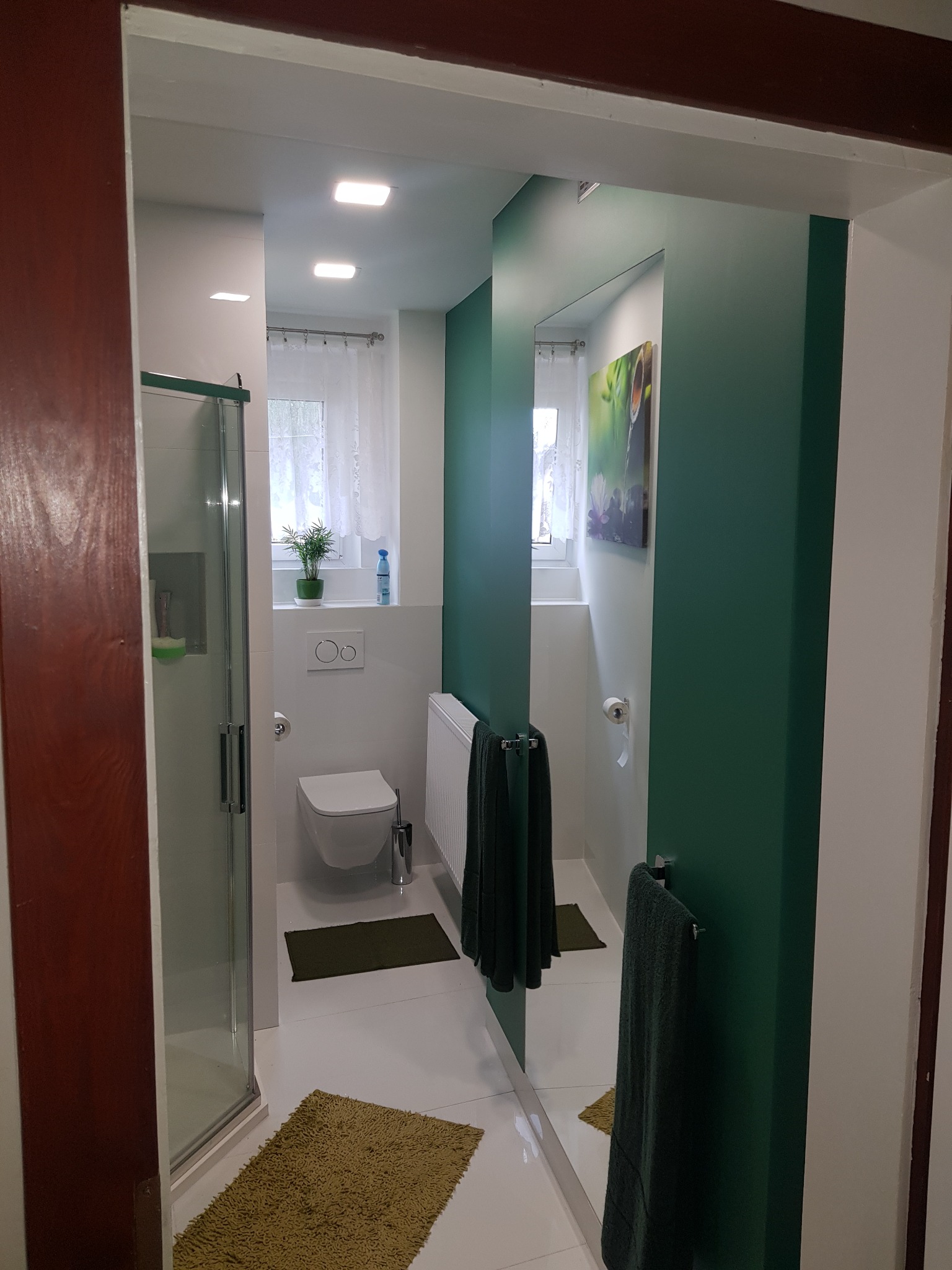 Nowoczesna łazienka z kabiną prysznicową, toaletą i umywalką, wykończona w bieli i butelkowej zieleni, z akcentami w postaci zielonych dywaników i ręczników, widziana z perspektywy wejścia.