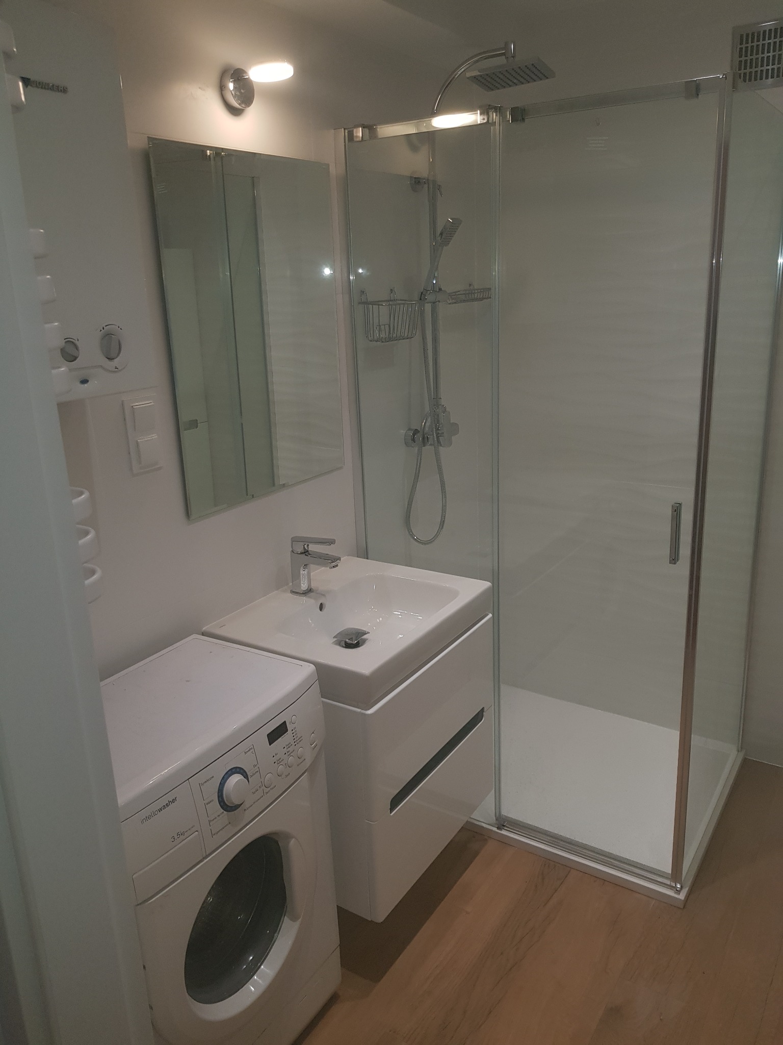 Nowoczesna, mała łazienka z kabiną prysznicową, umywalką z szafką i pralką, oświetlona punktowym światłem.