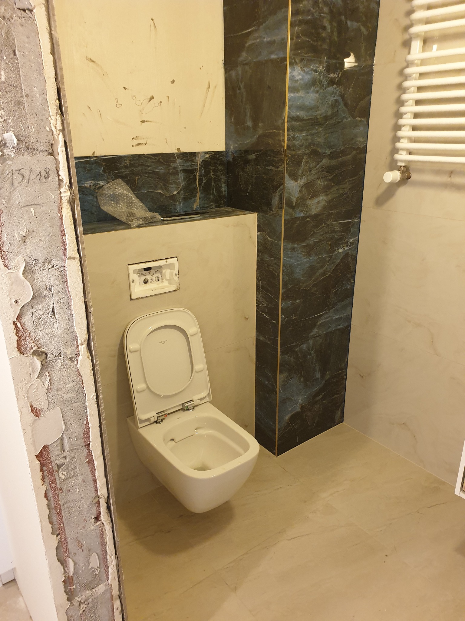 Wnętrze łazienki w trakcie montażu toalety podwieszanej, widoczne niedokończone obramowanie drzwi i fragment ściany z odchodzącą farbą, obok toalety położone płytki imitujące marmur, a na ścianie...