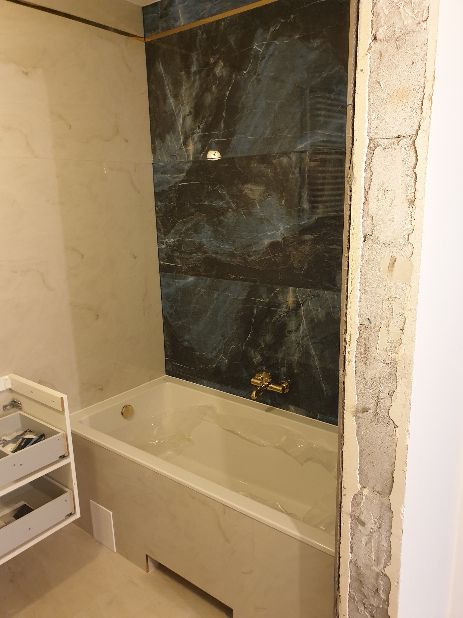 Wykończona łazienka z wanną obudowaną płytami imitującymi marmur, ściany wyłożone płytkami w dwóch kolorach z dekoracyjną listwą, widoczny fragment otwartej szafki i surowy framuga drzwi w trakcie...