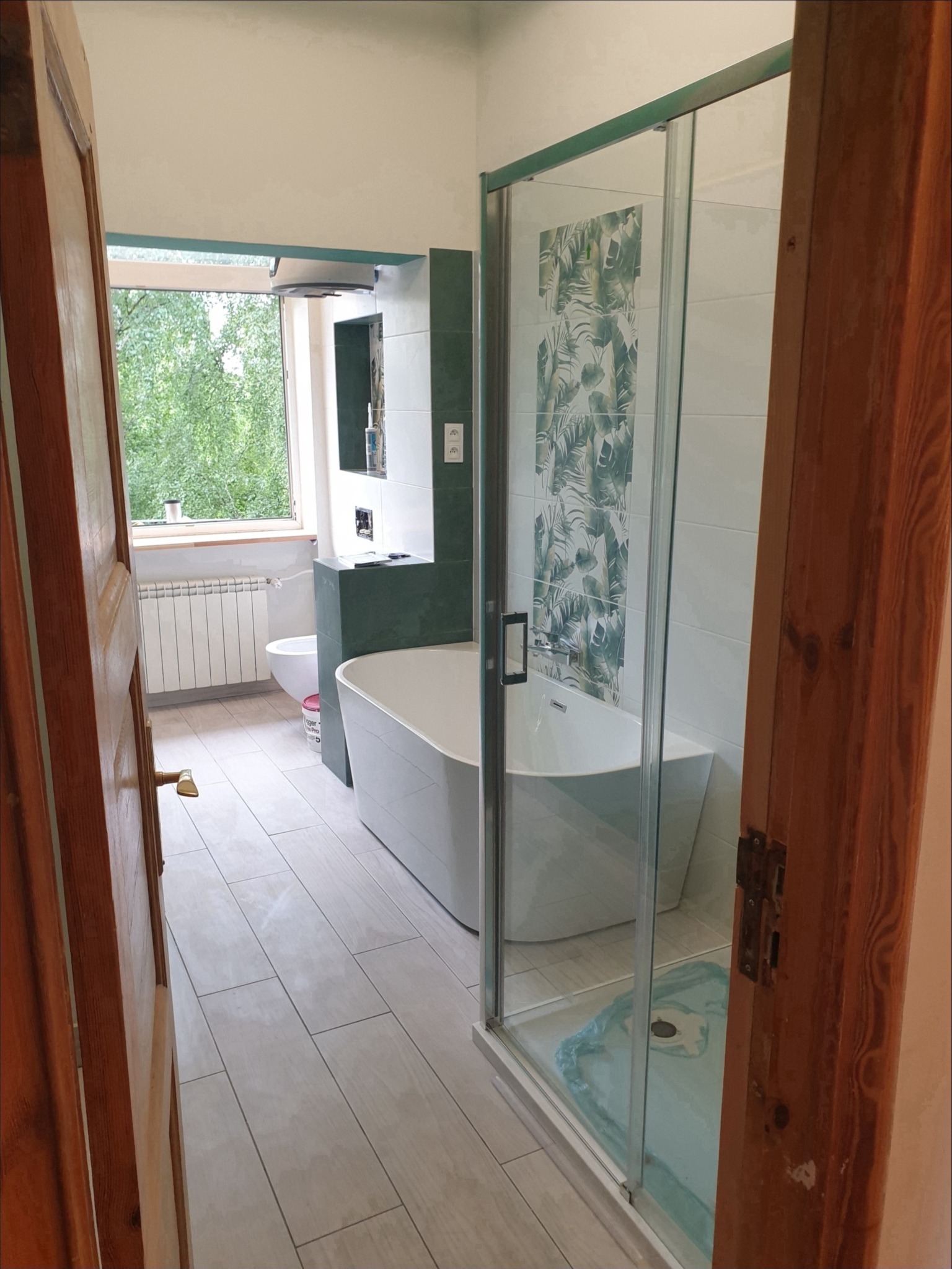 Wykończona łazienka z wolnostojącą wanną, kabiną prysznicową z motywem liści, drewnianymi drzwiami i oknem z widokiem na zieleń.