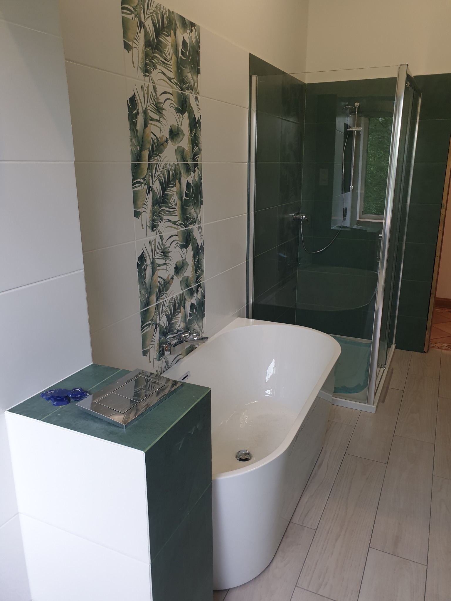 Nowoczesna łazienka z wolnostojącą wanną, kabiną prysznicową z ciemnozielonymi płytkami, biało-zielonymi płytkami z motywem tropikalnych liści oraz drewnopodobną podłogą z płytek.