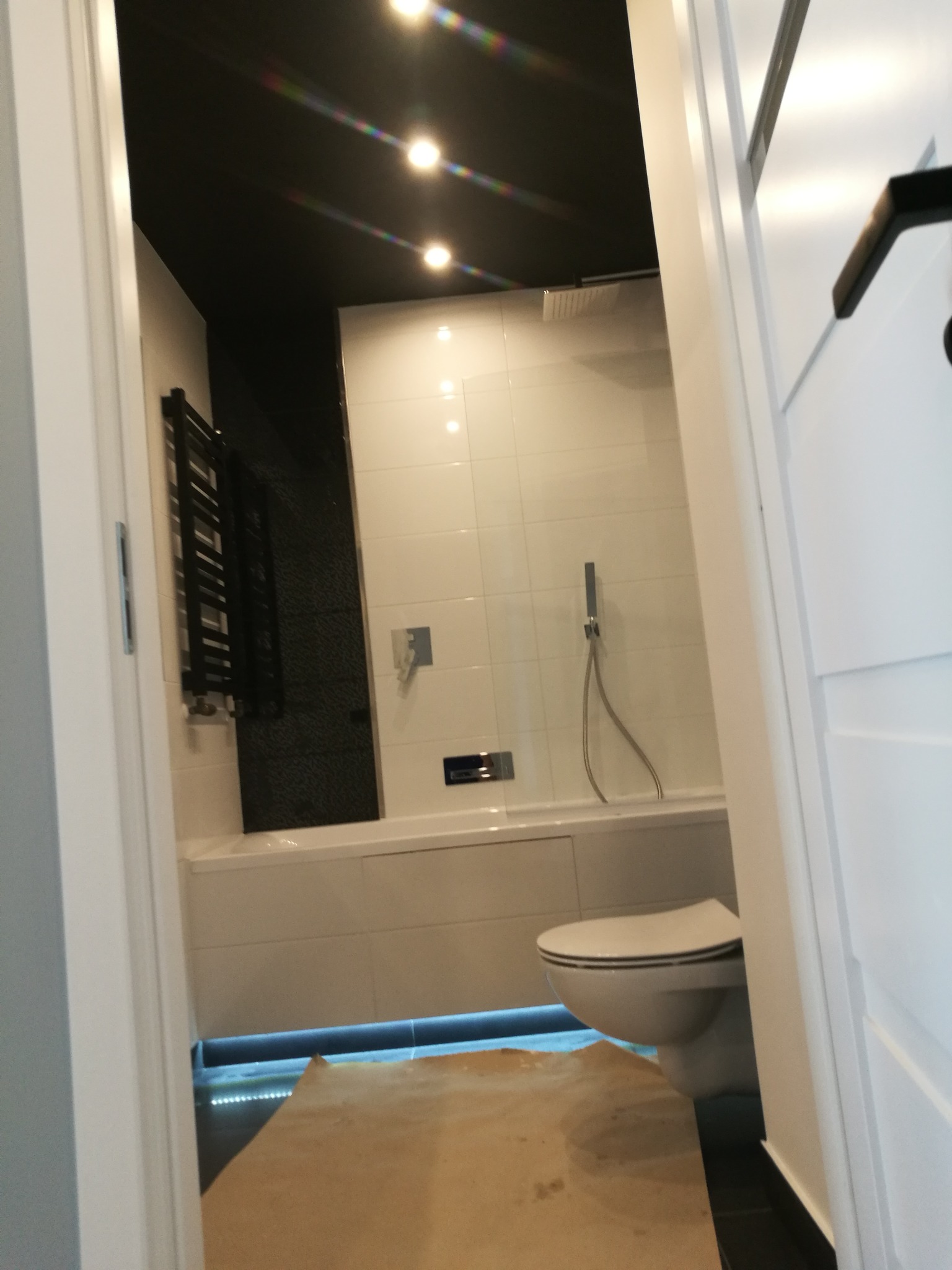 Nowoczesna łazienka z białymi płytkami, podświetlaną wanną, czarnym sufitem i grzejnikiem drabinkowym, widok zza uchylonych drzwi.