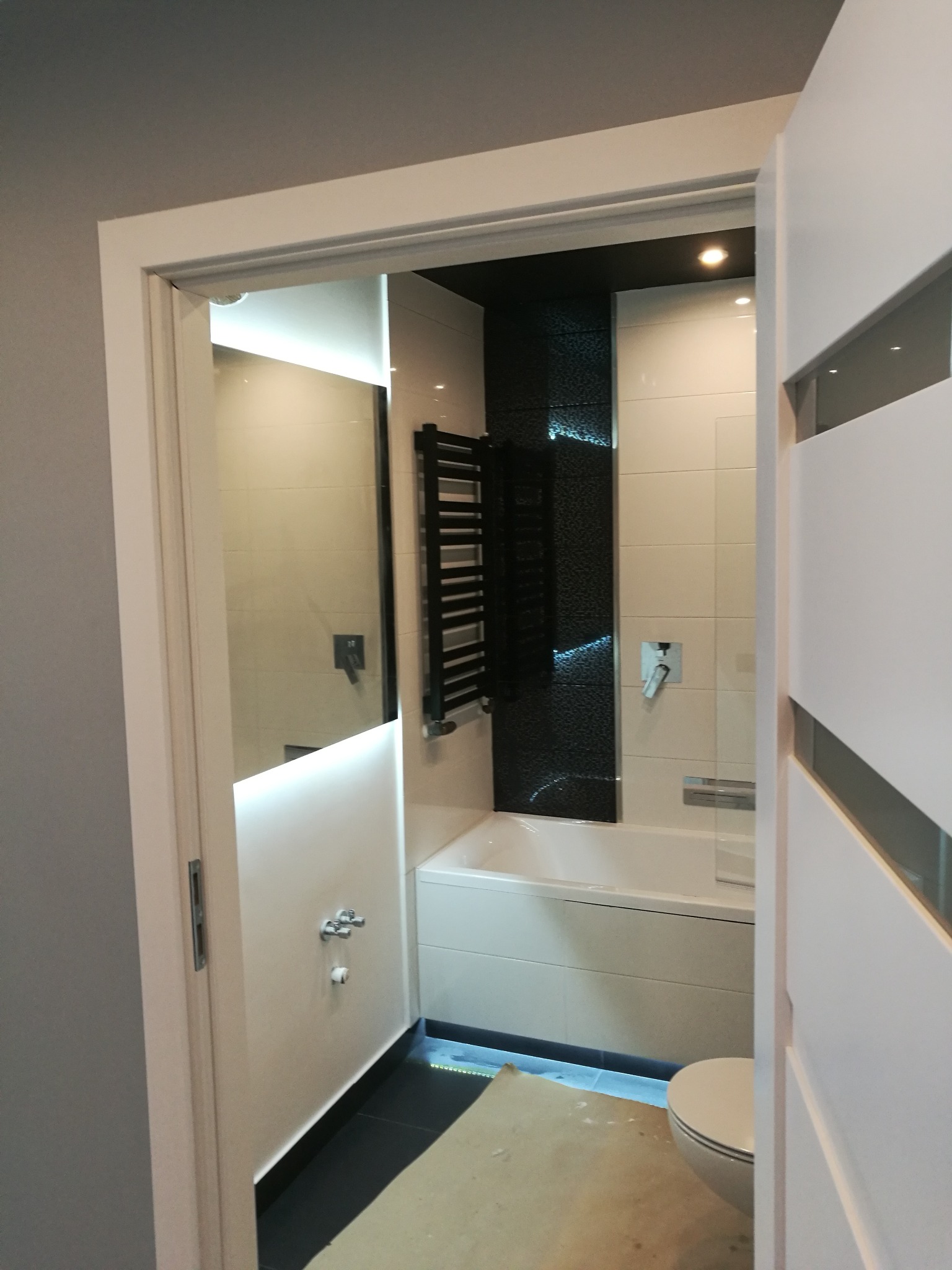 Nowoczesna łazienka z czarno-białymi płytkami, podświetlanym lustrem i grzejnikiem drabinkowym, widok zza uchylonych drzwi z widocznym fragmentem niezabezpieczonej podłogi.