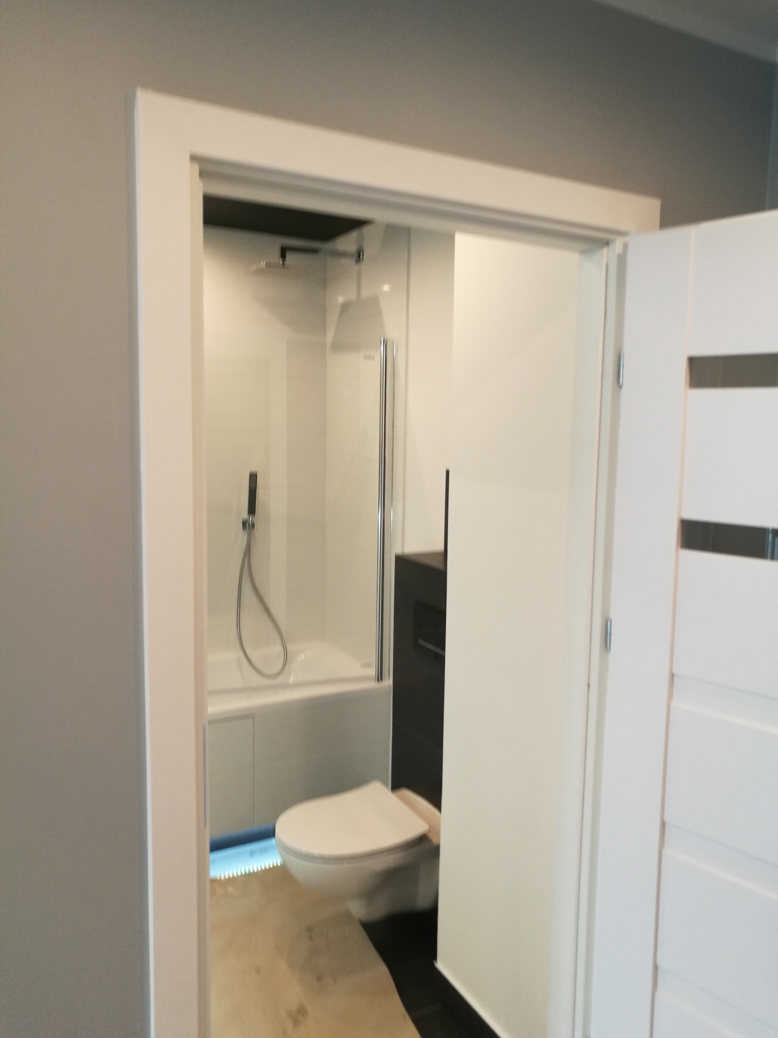 Nowoczesna, minimalistyczna łazienka z kabiną prysznicową nad wanną, wiszącą toaletą i ciemną szafką pod umywalką, widoczna przez białe, częściowo otwarte drzwi.