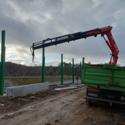 Kings - Montaż prefabrykowanych betonowych elementów ogrodzenia przy drodze ekspresowej za pomocą dźwigu samochodowego, widoczne zielone słupki konstrukcyjne