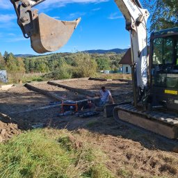 Kings - Bobcat kopie rów pod fundamenty na działce z widokiem na górzysty krajobraz, pracownicy używają poziomicy i agregatu prądotwórczego.