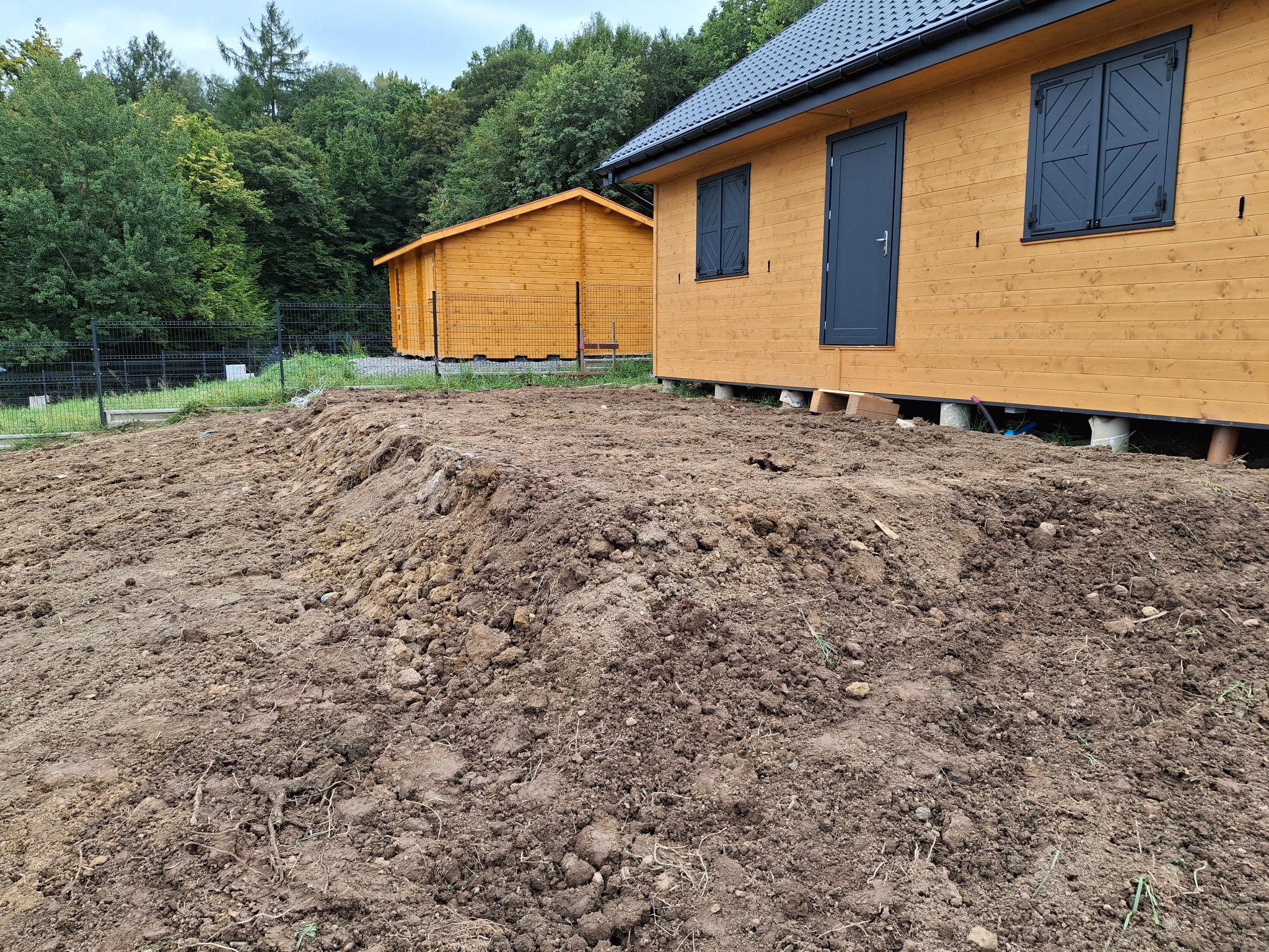 Dwa drewniane domki letniskowe na podwyższeniu, widoczne świeżo usypane ziemne podłoże przed budynkami, w tle zalesiony teren.