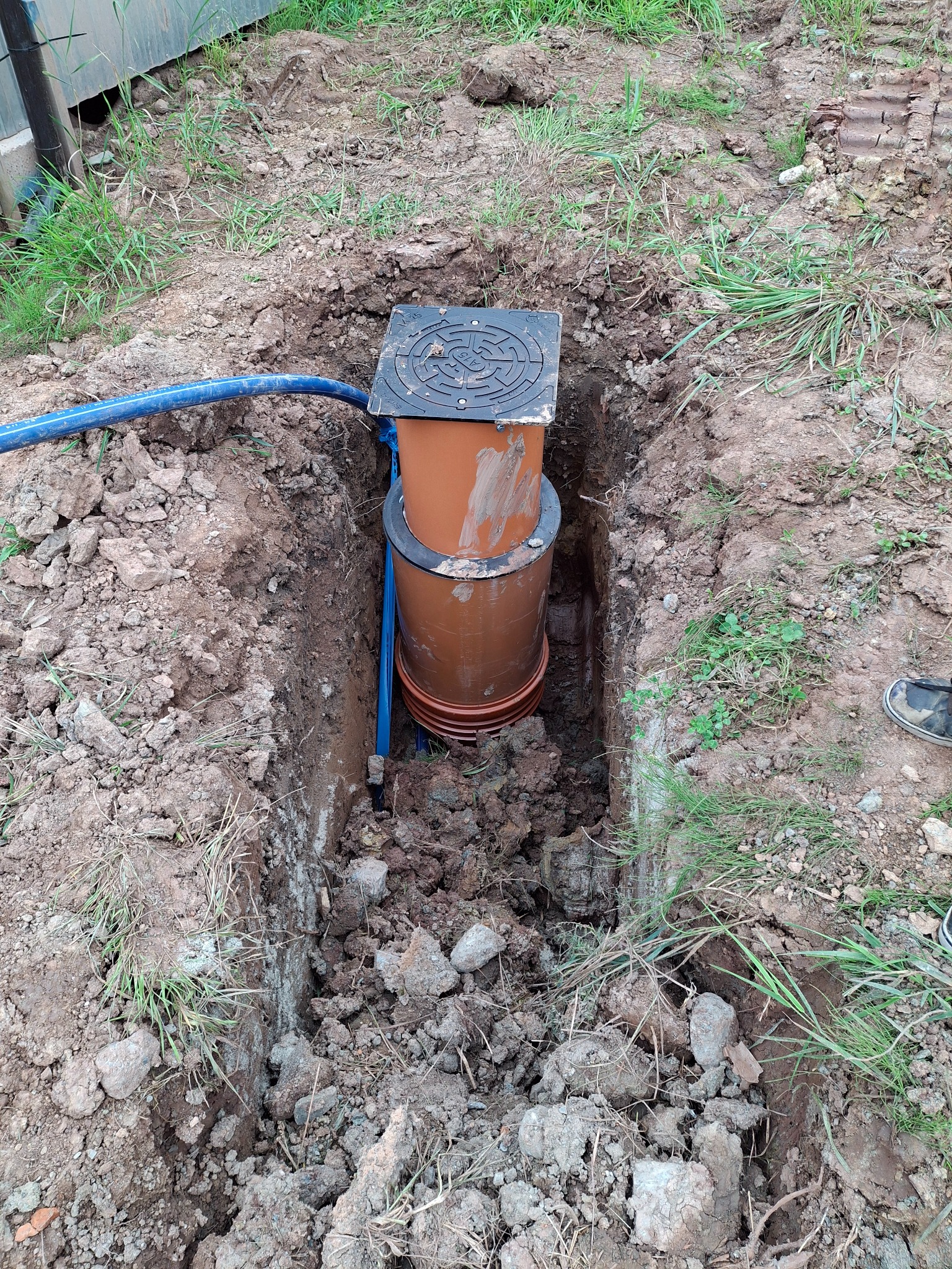 Głęboki wykop w ziemi z widoczną studzienką kanalizacyjną z rur PCV i niebieskim przewodem, otoczoną gruzem i trawą.