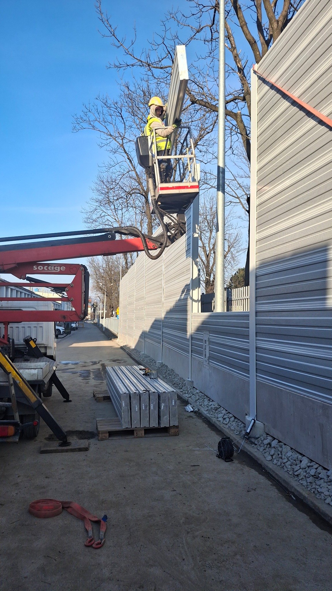 Montaż paneli akustycznych przy drodze przez pracownika w podnośniku koszowym, widoczne panele na palecie i fragment żurawia.