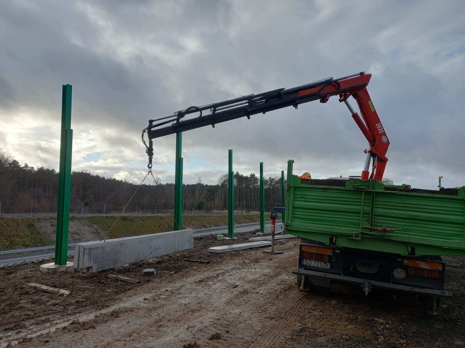 Montaż prefabrykowanych betonowych elementów ogrodzenia przy drodze ekspresowej za pomocą dźwigu samochodowego, widoczne zielone słupki konstrukcyjne