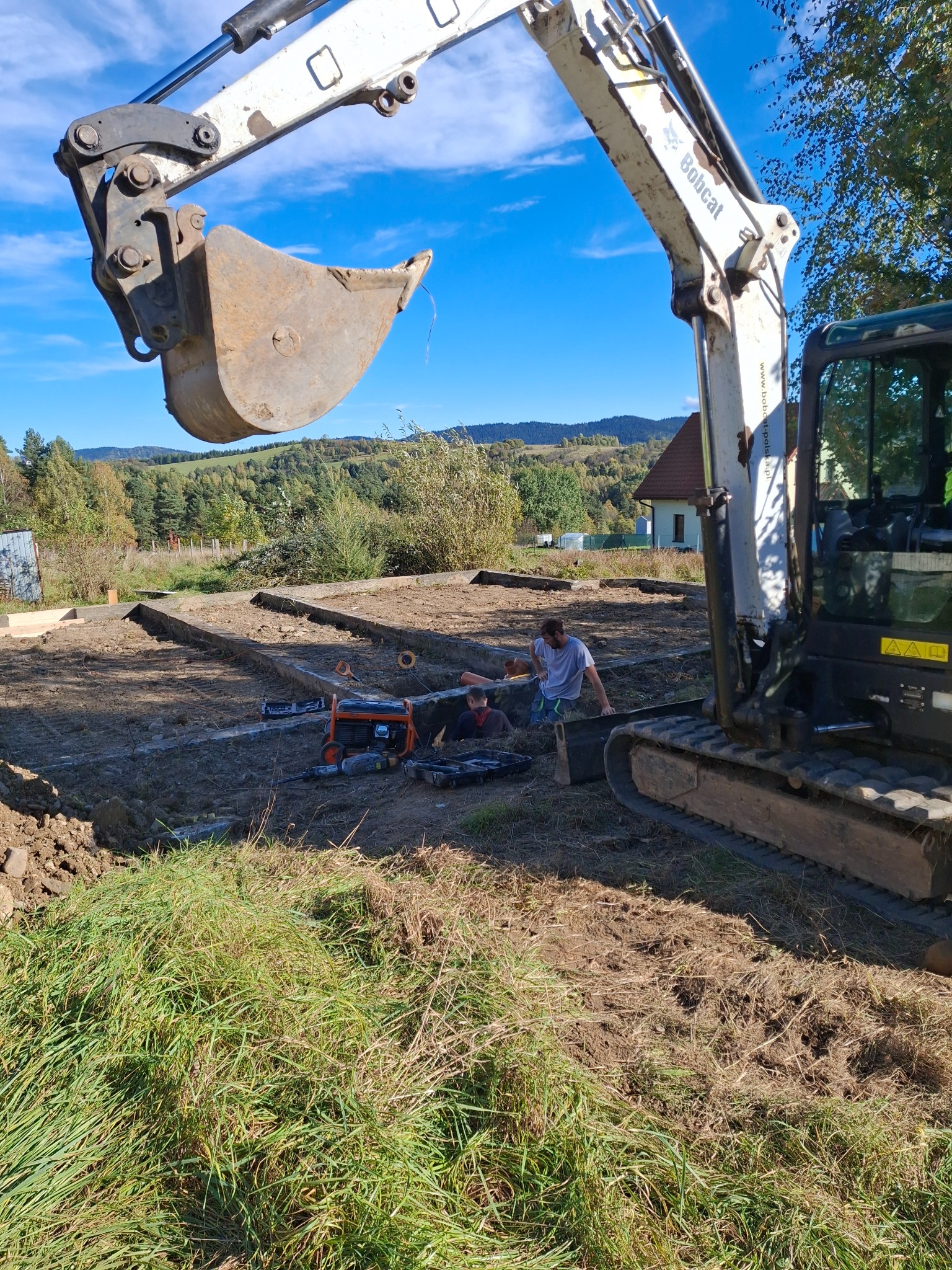 Bobcat kopie rów pod fundamenty na działce z widokiem na górzysty krajobraz, pracownicy używają poziomicy i agregatu prądotwórczego.
