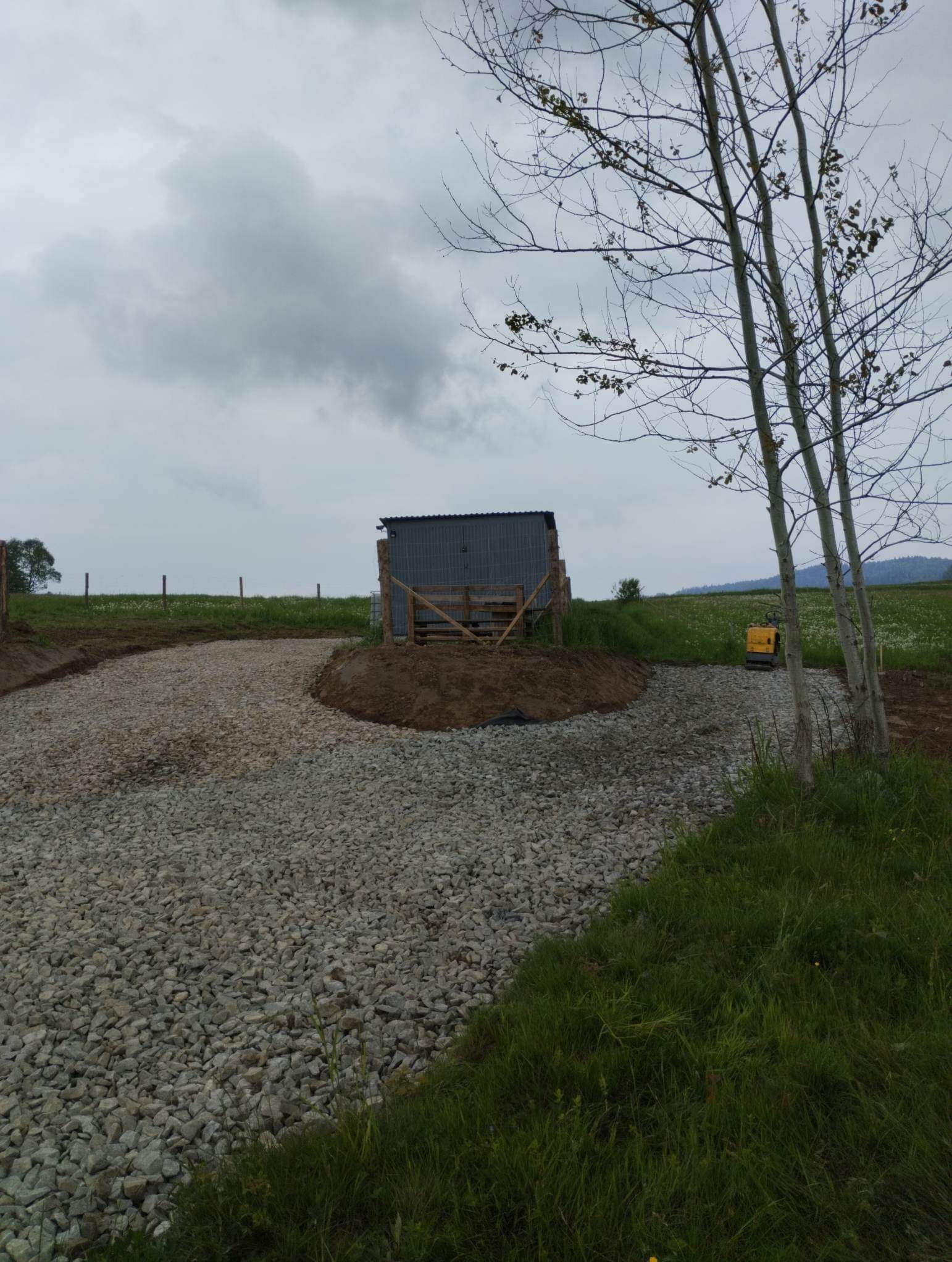 Teren wyrównany żwirem z małą koparką w tle, obok rosną drzewa bez liści, a w oddali widać budynek gospodarczy.