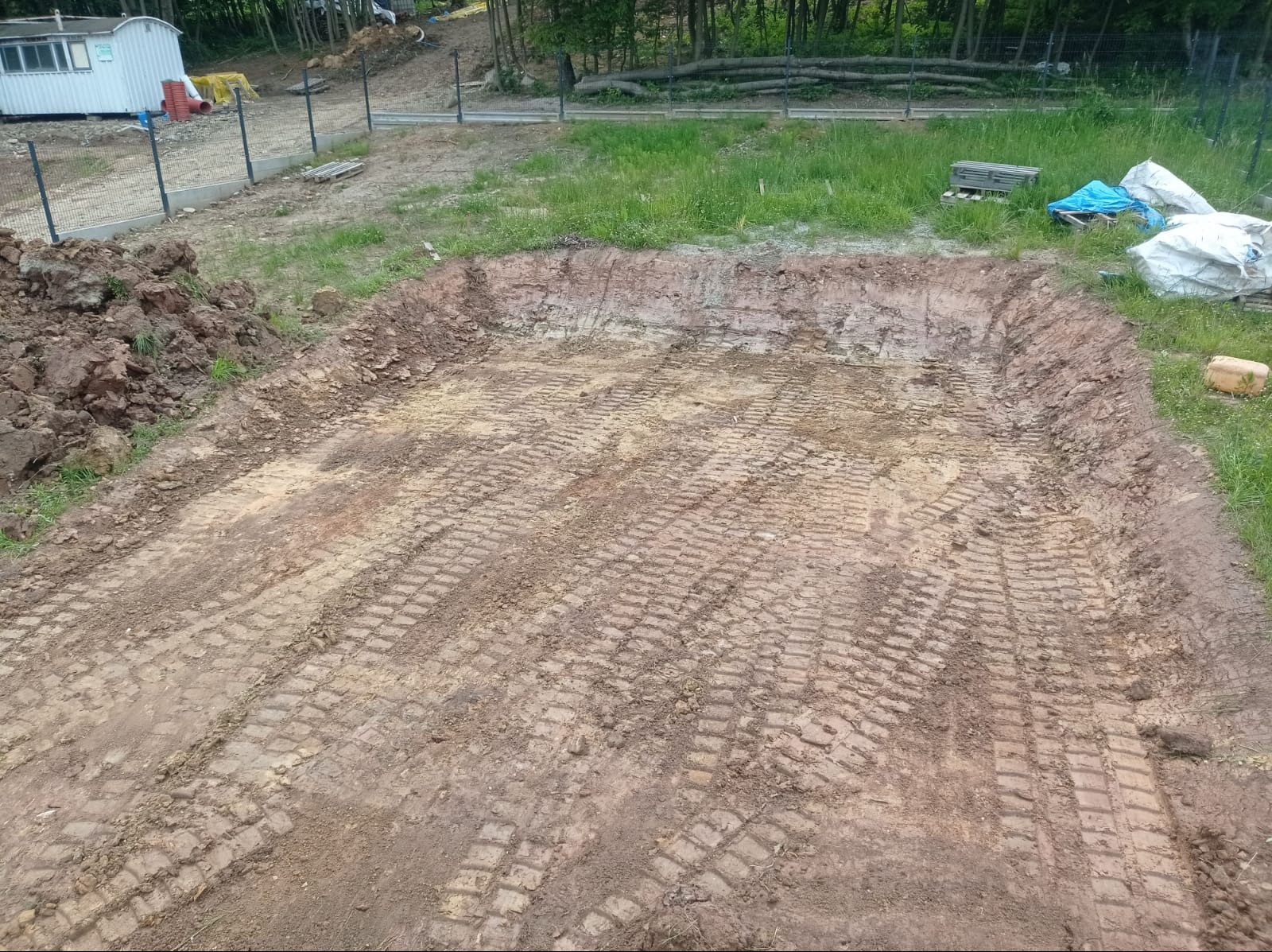 Wykop ziemny w kształcie prostokąta z wyraźnymi śladami opon ciężkiego sprzętu na dnie, obok sterta ziemi i częściowo ogrodzony teren budowy z zalesionym tłem.