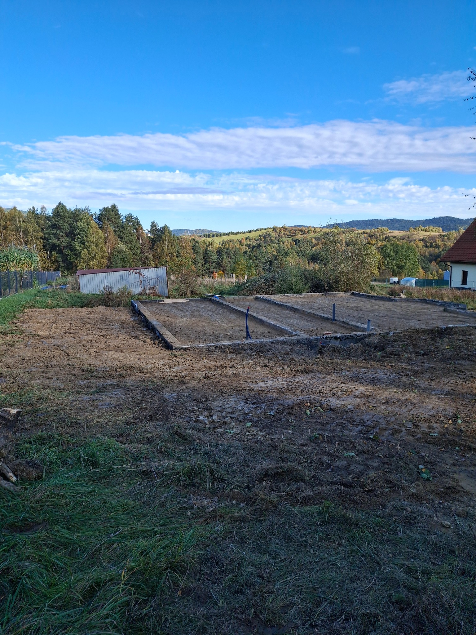 Przygotowany teren pod budowę z wylanymi fundamentami w postaci betonowych belek, widoczne wystające rury, w tle budynek mieszkalny i zalesione wzgórza pod błękitnym niebem.