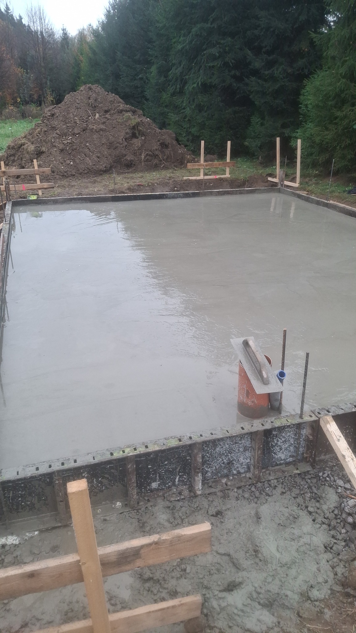 Świeżo wylany beton na przygotowanym fundamencie, widoczne szalunki, narzędzia budowlane i sterta ziemi w tle.