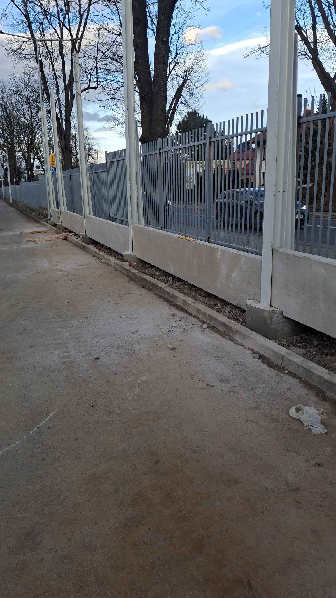 Betonowy fundament ogrodzenia z metalowymi słupkami i przęsłami, widoczny z poziomu chodnika, z fragmentami cegieł i gruzu w szczelinie przy krawężniku.