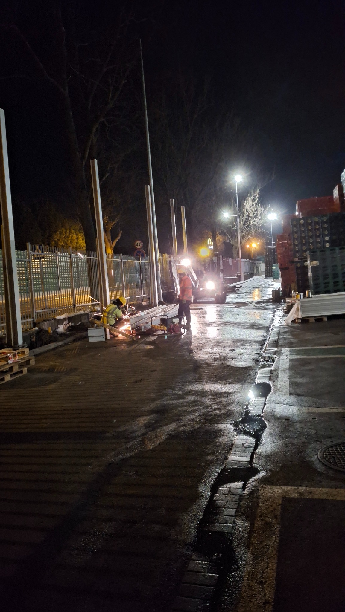 Prace spawalnicze przy montażu słupów oświetleniowych w nocy; pracownicy w odblaskowych kamizelkach, widoczny sprzęt budowlany i palety z materiałami.