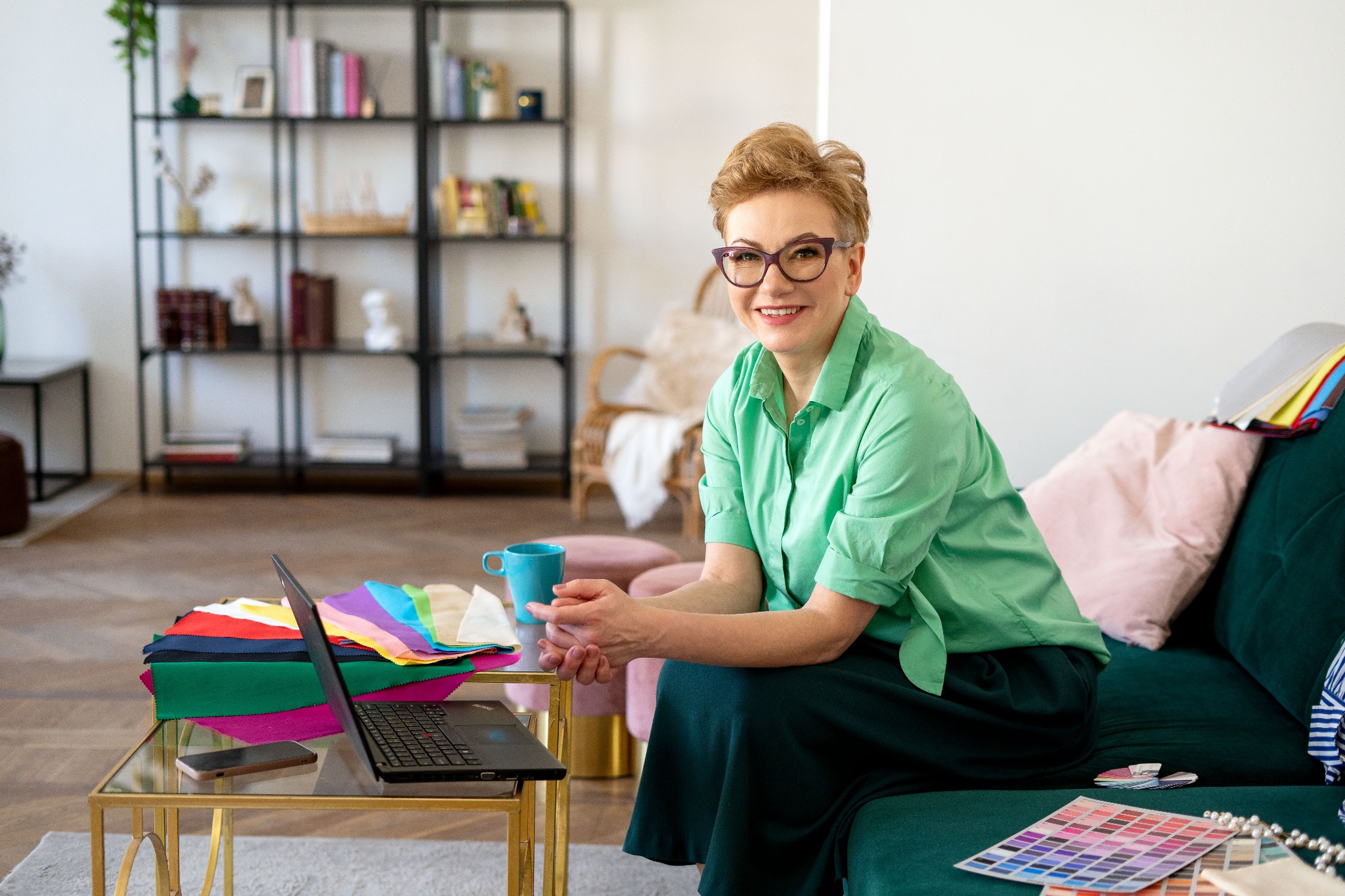 Uśmiechnięta kobieta w okularach siedzi na zielonej sofie, obok stolika z laptopem, próbkami materiałów i kubkiem; w tle regał z książkami.