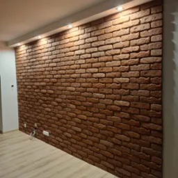Ściana wykończona czerwoną cegłą dekoracyjną, oświetlona punktowymi lampami sufitowymi, z widocznymi gniazdkami elektrycznymi u dołu i jasną podłogą panelową.