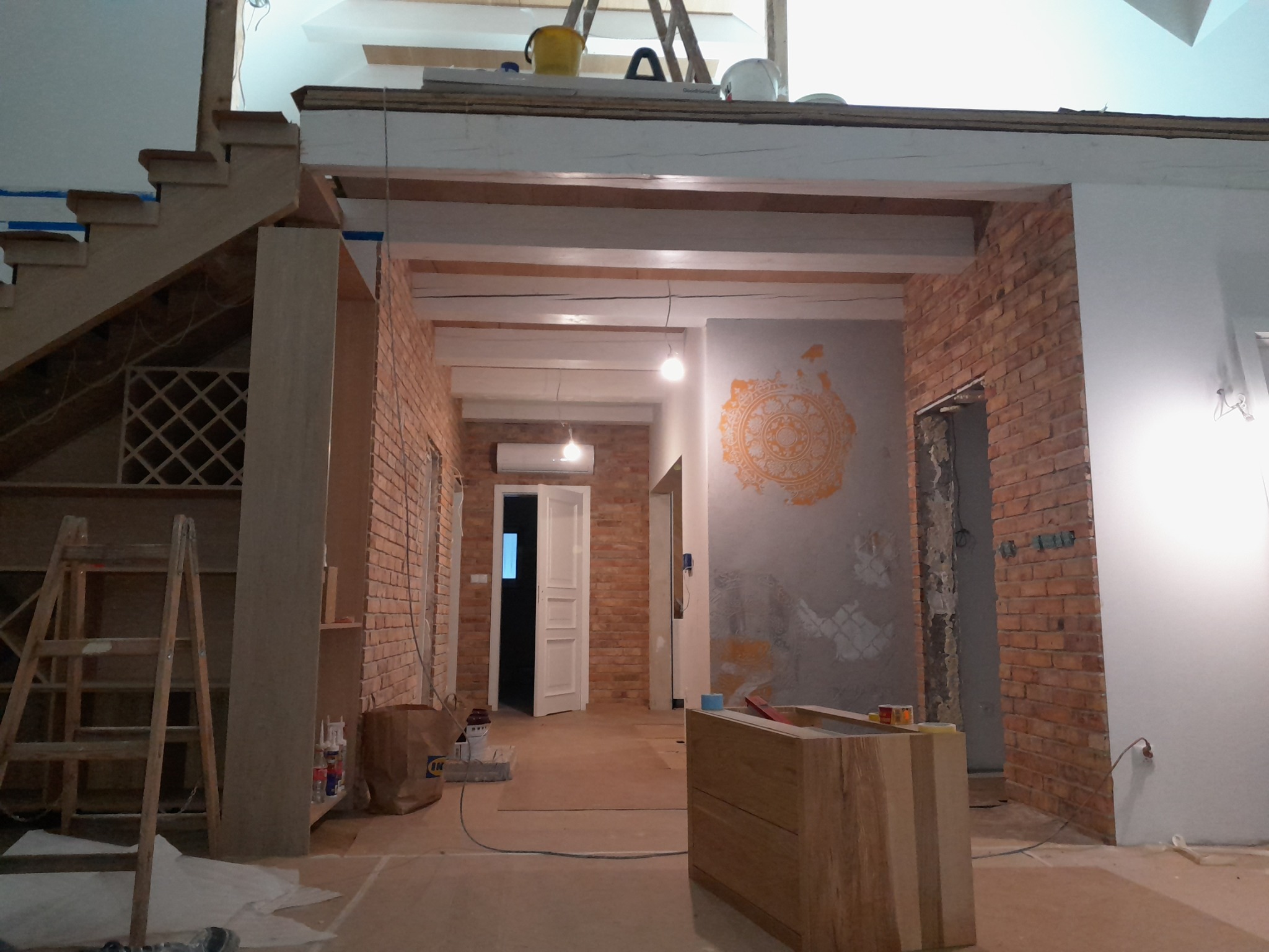 Wnętrze w trakcie remontu: widoczne odsłonięte cegły, surowe drewniane belki stropowe, schody w trakcie wykańczania, drabina oparta o ścianę, dekoracyjny wzór na ścianie, podłoga zabezpieczona...