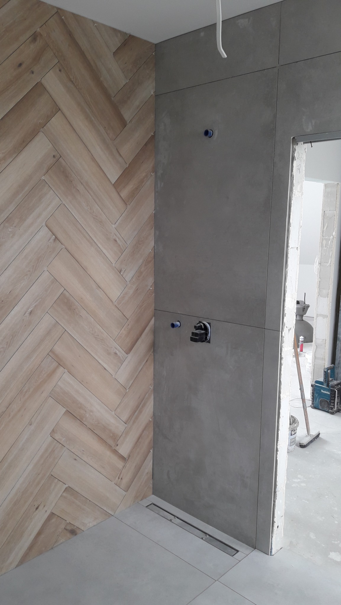 Ściana łazienki wykończona płytkami drewnopodobnymi ułożonymi w jodełkę, sąsiadująca z szarą ścianą z ukrytą instalacją wodną i odpływem liniowym w podłodze, widoczny sprzęt budowlany w tle.