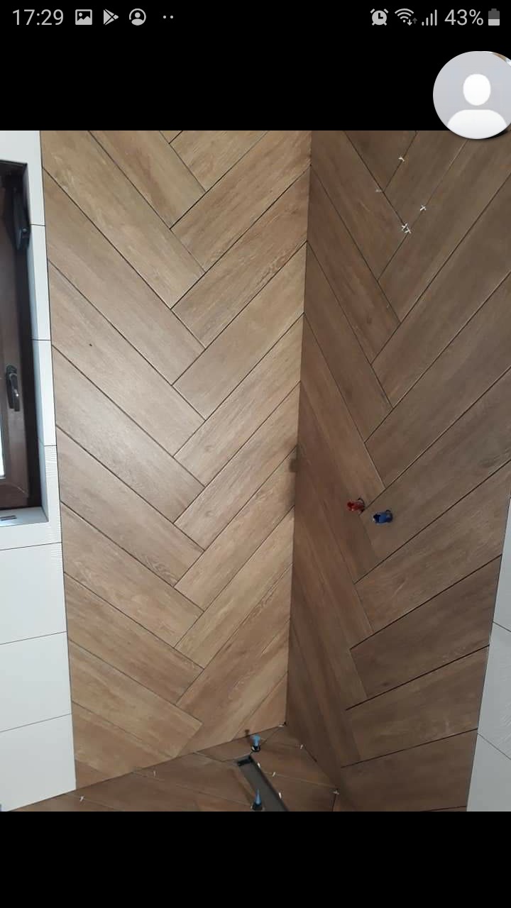 Ukończona ściana w łazience z płytek imitujących drewno, ułożonych w jodełkę, widoczne elementy instalacji wodnej i fragment okna.