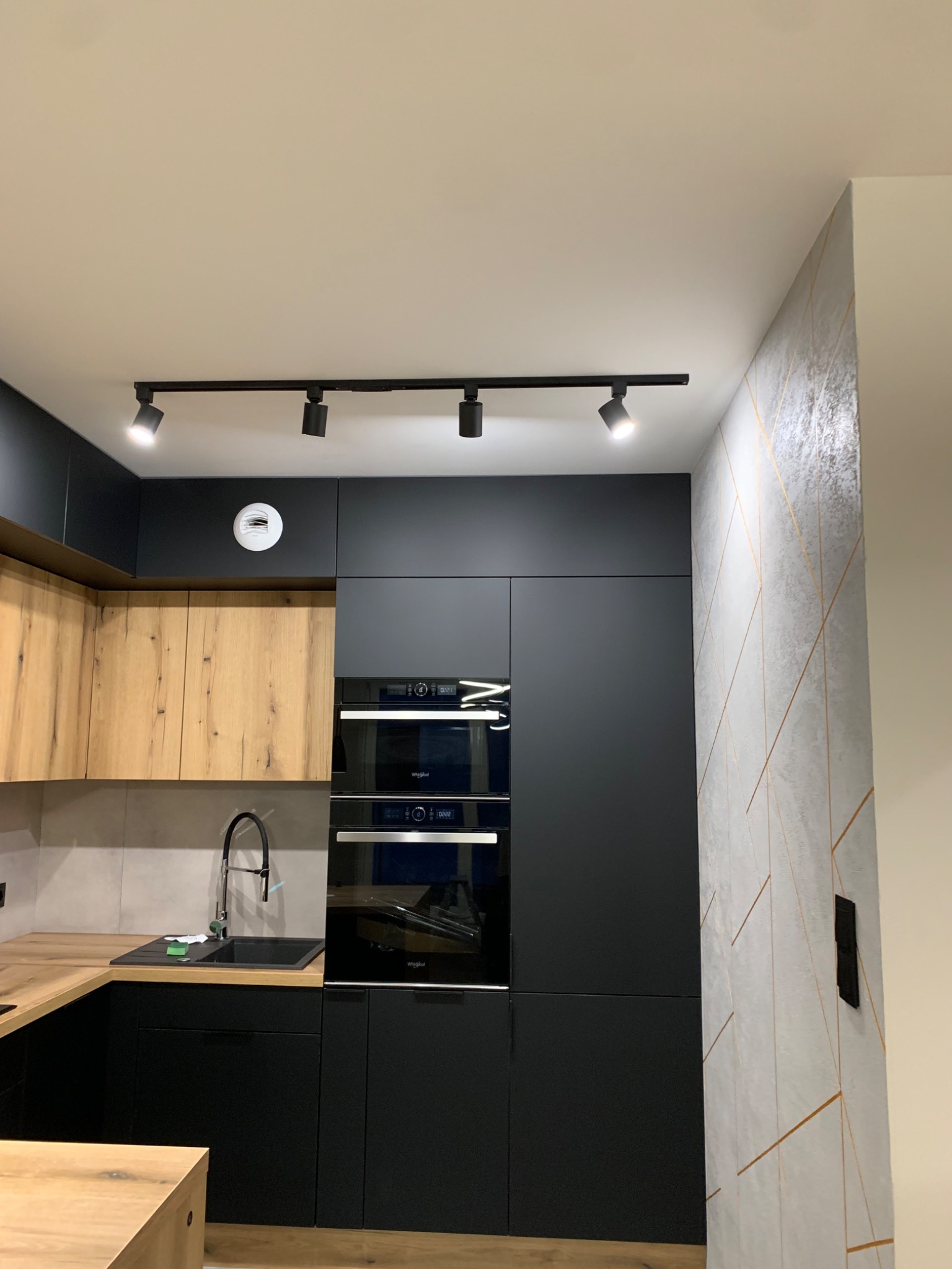 Nowoczesna kuchnia z czarnymi i drewnianymi szafkami, oświetleniem szynowym i dekoracyjną ścianą z geometrycznym wzorem.