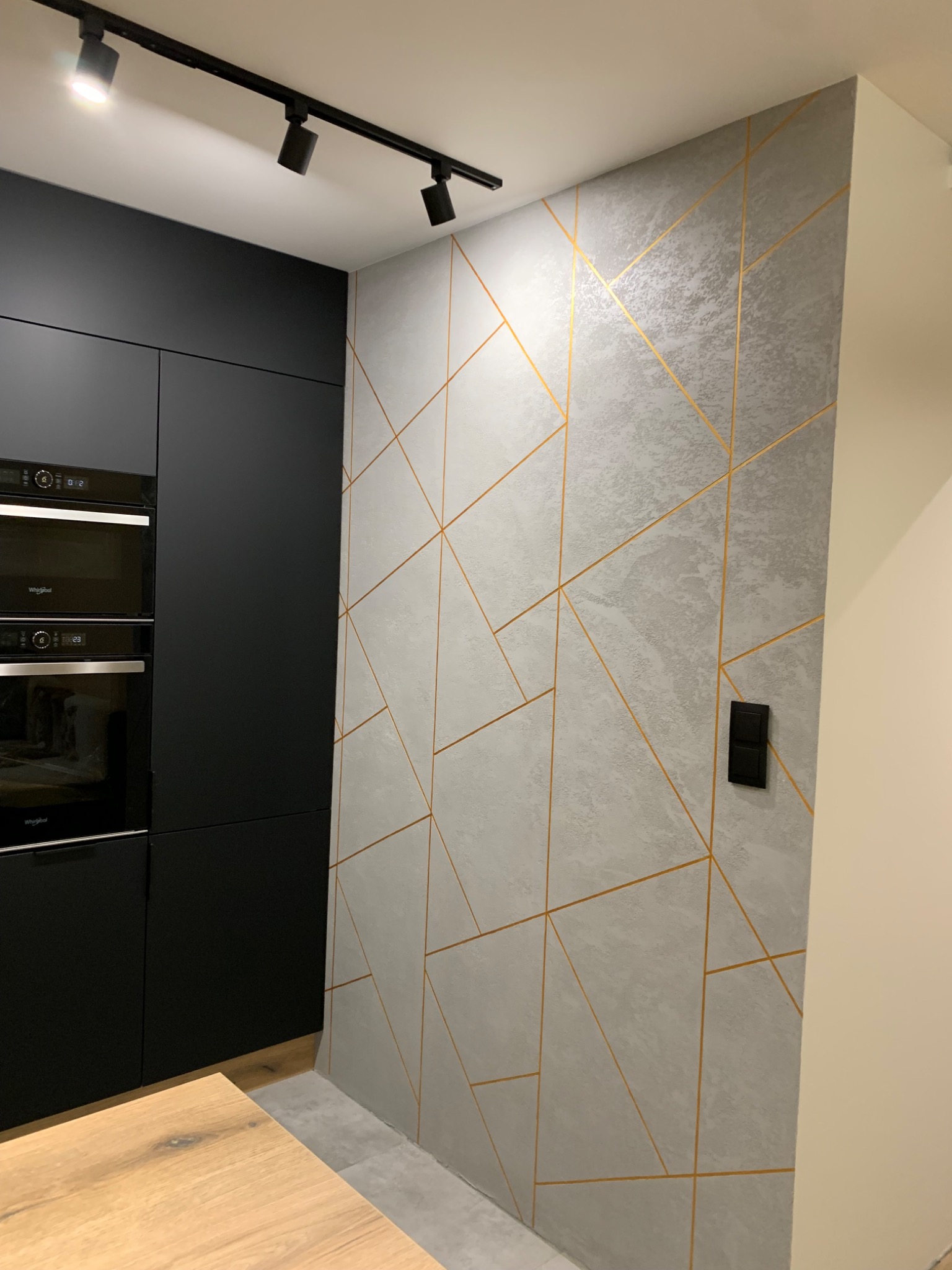 Ściana w kuchni pokryta szarą gładzią z geometrycznym wzorem w złotym kolorze, obok zabudowa kuchenna w czarnym macie i drewniany blat.