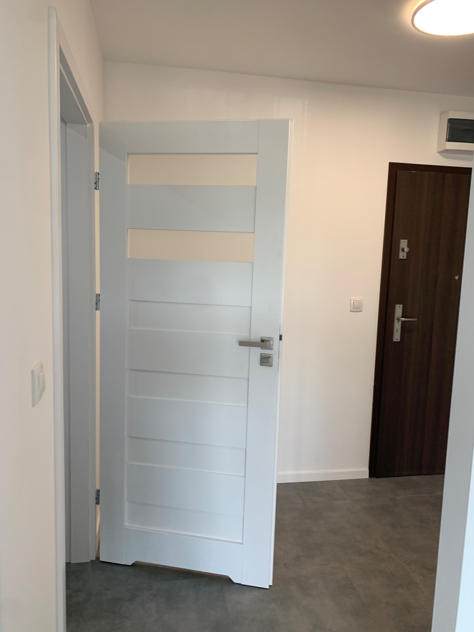 Białe drzwi wewnętrzne z poziomymi przeszkleniami, uchwyt ze stali nierdzewnej, widoczne zawiasy, szara podłoga, ciemnobrązowe drzwi wejściowe w tle.