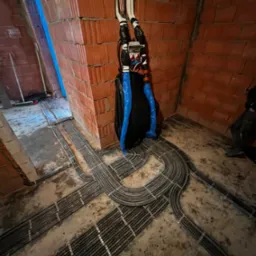 Rozprowadzenie kabli elektrycznych w peszlach, mocowanych do podłoża w surowym stanie, widok na instalację w trakcie realizacji w pomieszczeniu z ceglanymi ścianami.