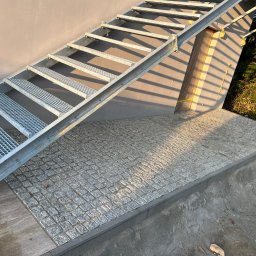 Usługi Brukarsko-Ogrodzeniowe UZBUD Marcin Guz - Montaż schodów i kostki granitowej 8x8 w Pruszczu Gdańskim. Widoczne metalowe schody prowadzące na podest wyłożony kostką. Słoneczne oświetlenie podkreśla fakturę.