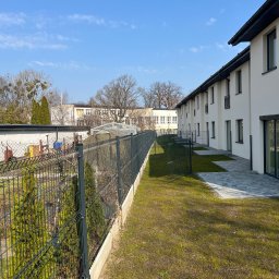 Usługi Brukarsko-Ogrodzeniowe UZBUD Marcin Guz - Nowoczesne ogrodzenie panelowe w kolorze grafitowym, oddzielające posesje szeregowych domów z kostką brukową i trawnikiem. Błękitne niebo w tle.